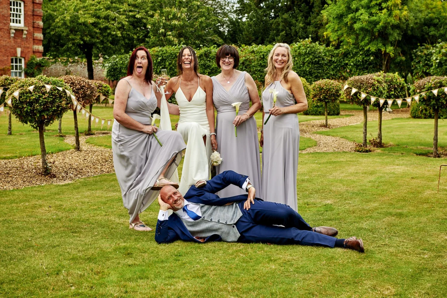 Groom lies on garden grass as bride and bridesmaids stand behind him.