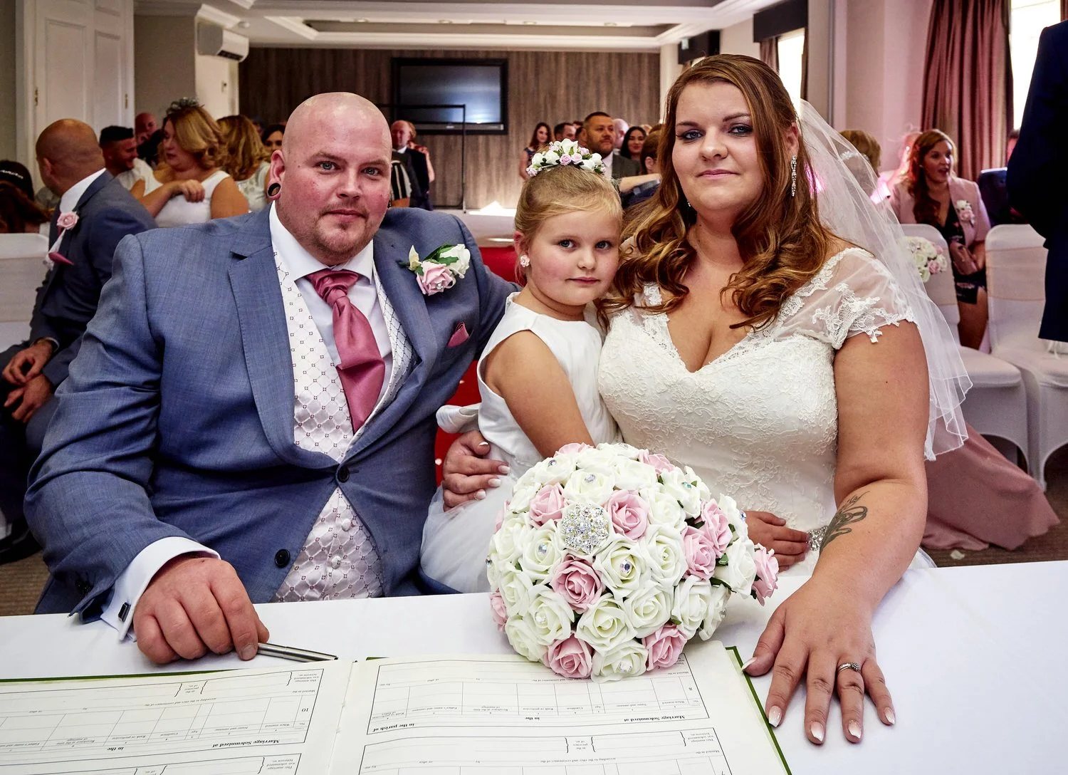 The bride and groom sign the register with their guests sat behind them, young bridesmaid is on the bride’s lap.