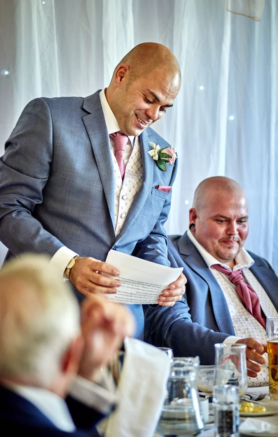 The best man reads a speech standing next to the groom who is smiling.