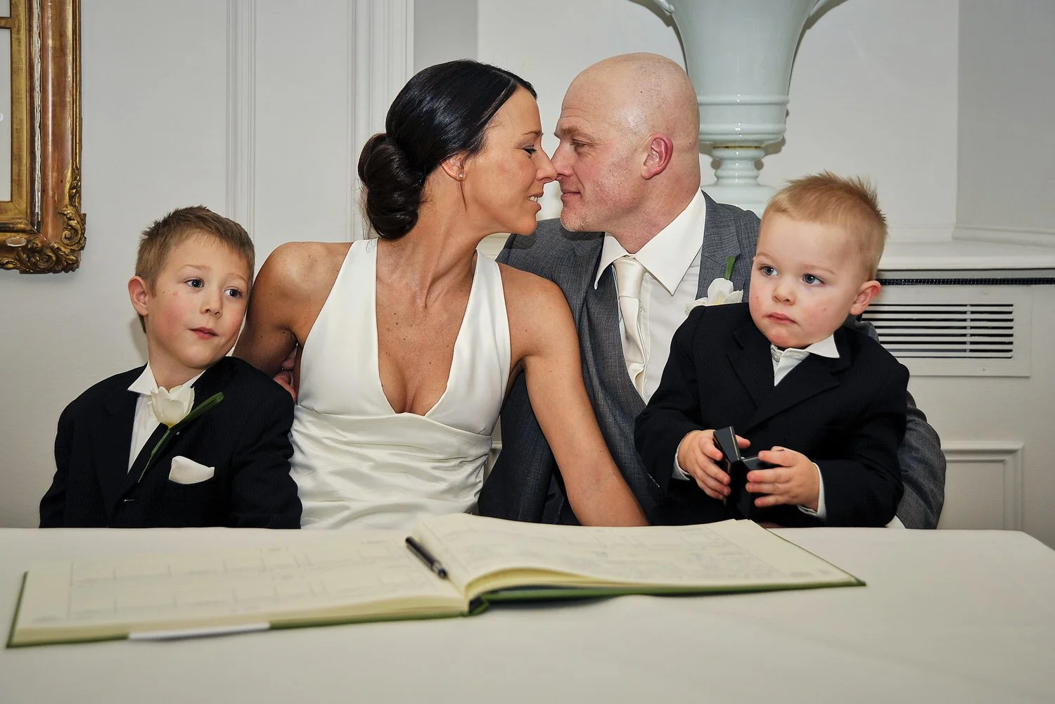 Bride and groom kiss while signing register with two small children on their laps.