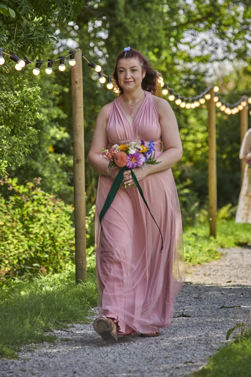Bridesmaid walks down a tree-lined path holding flowers as she joins the wedding party.