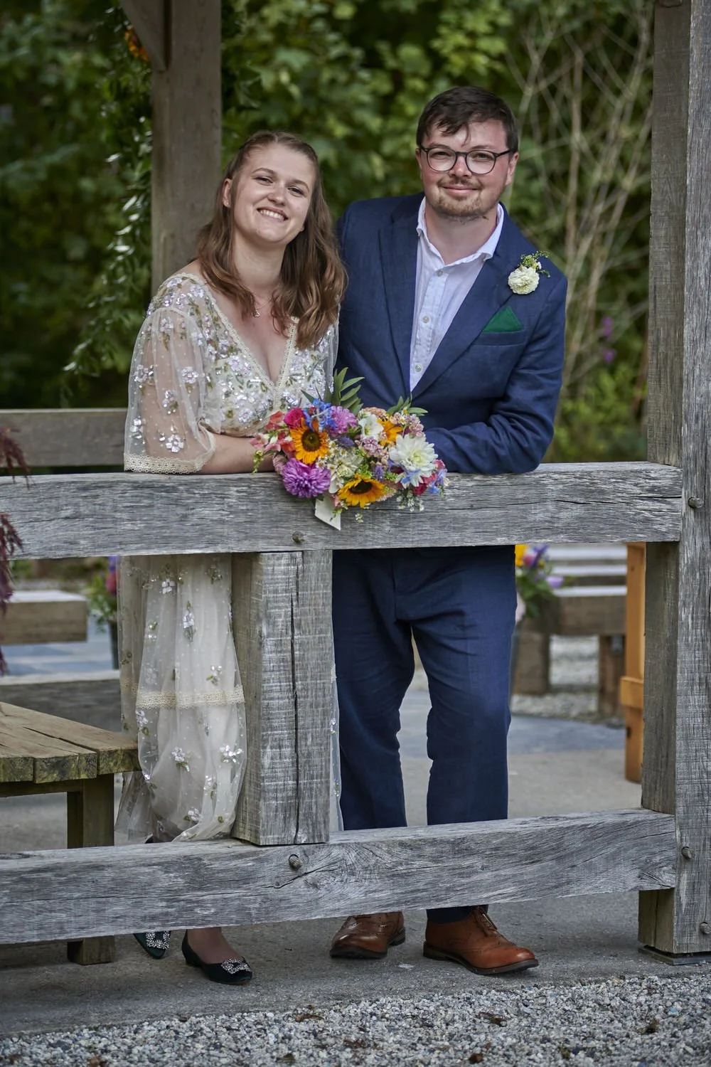 Bride and groom lean on a wooden fence and smile at the camera.