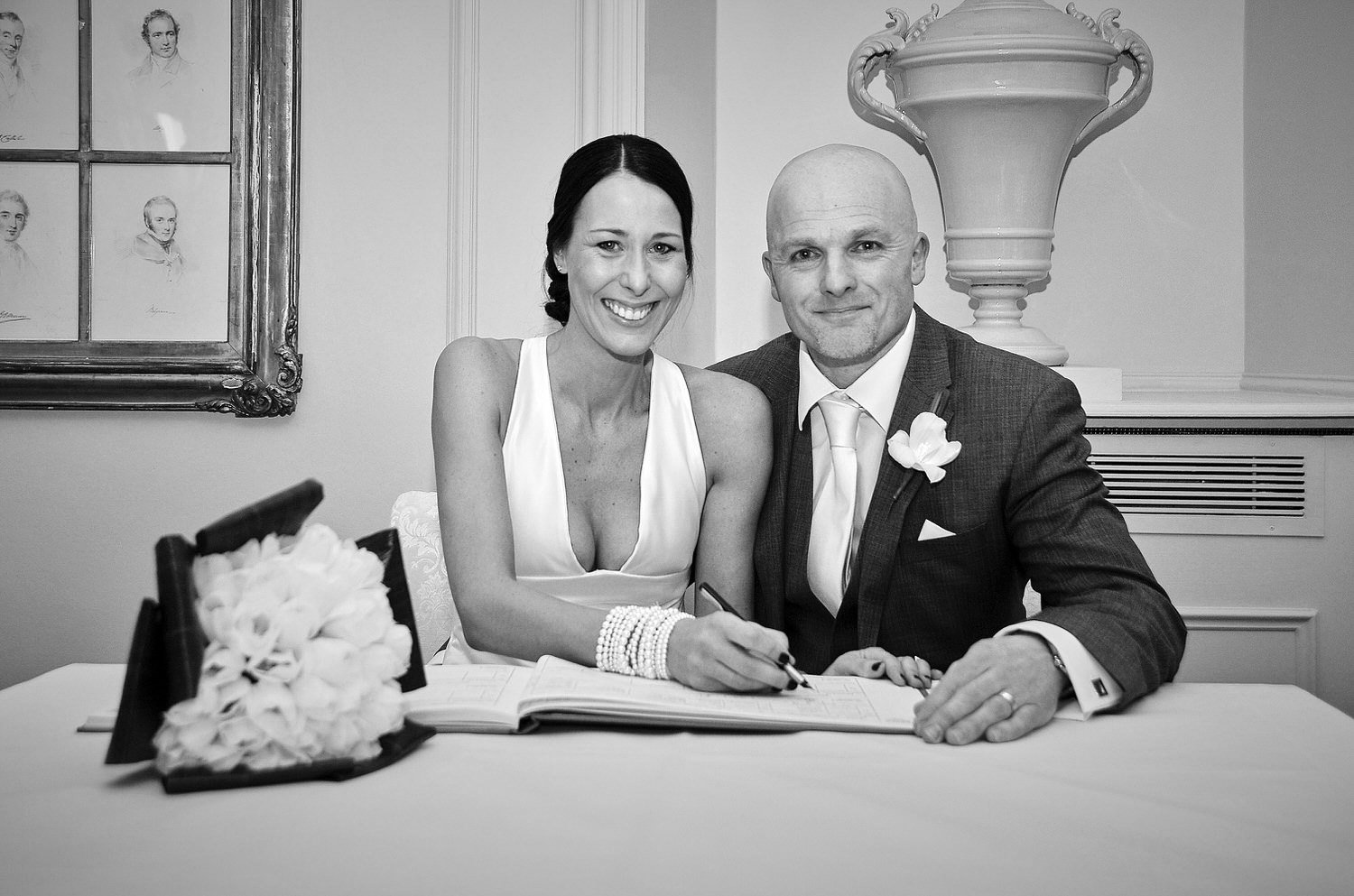 Bride and groom signing the register smile at camera – black and white image.