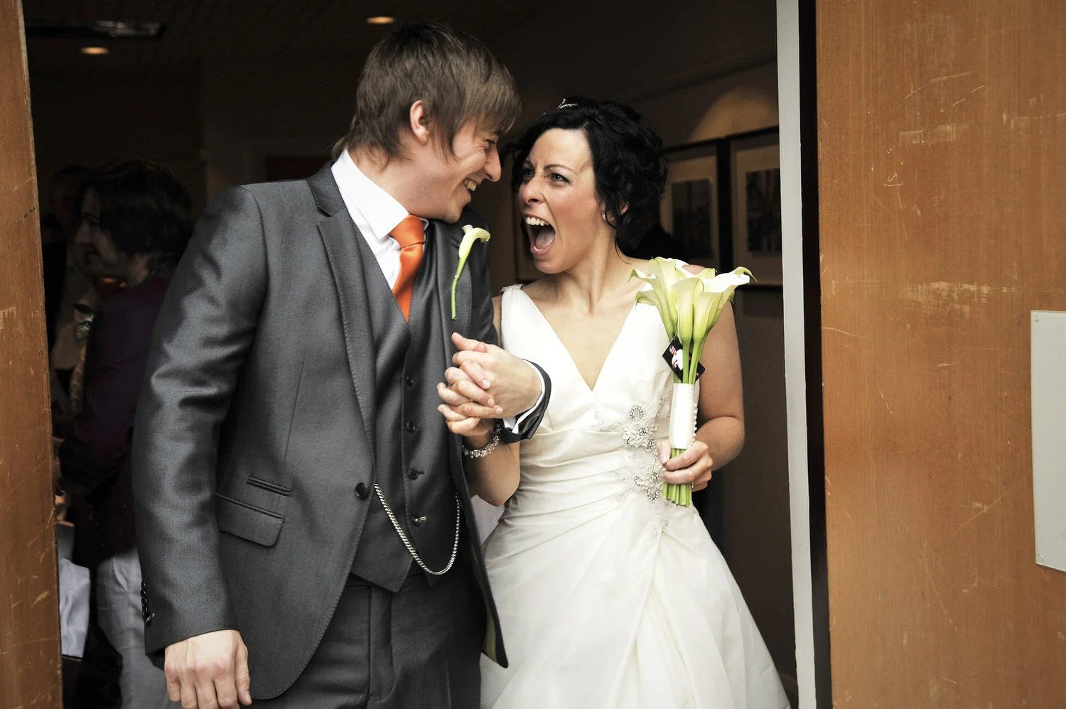 Bride and groom walk out of a door holding hands and grinning at each other.