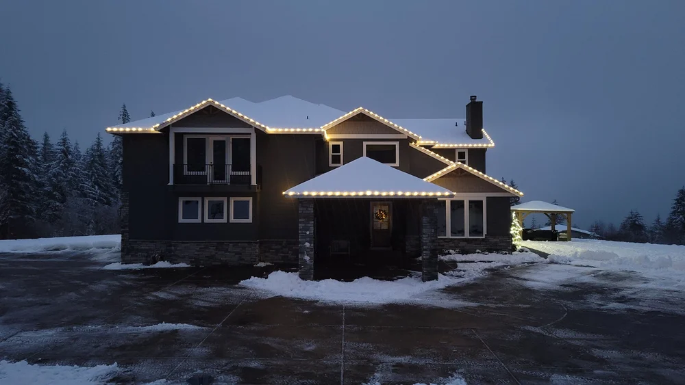 Large warm white bulbs evenly spaced along the rooflines of a snowy two-story home