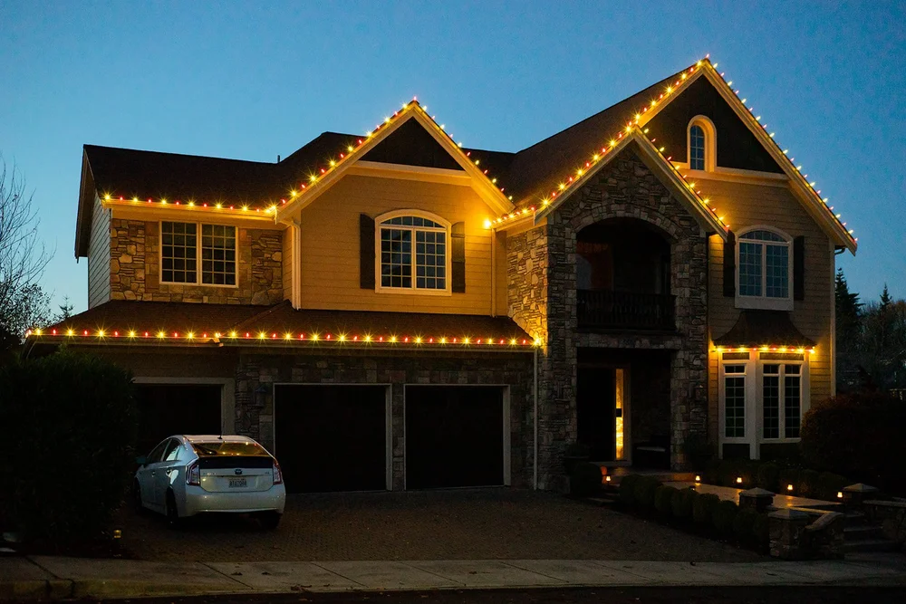 Two-story stone home with warm white C9 Christmas lights along every roofline at dusk