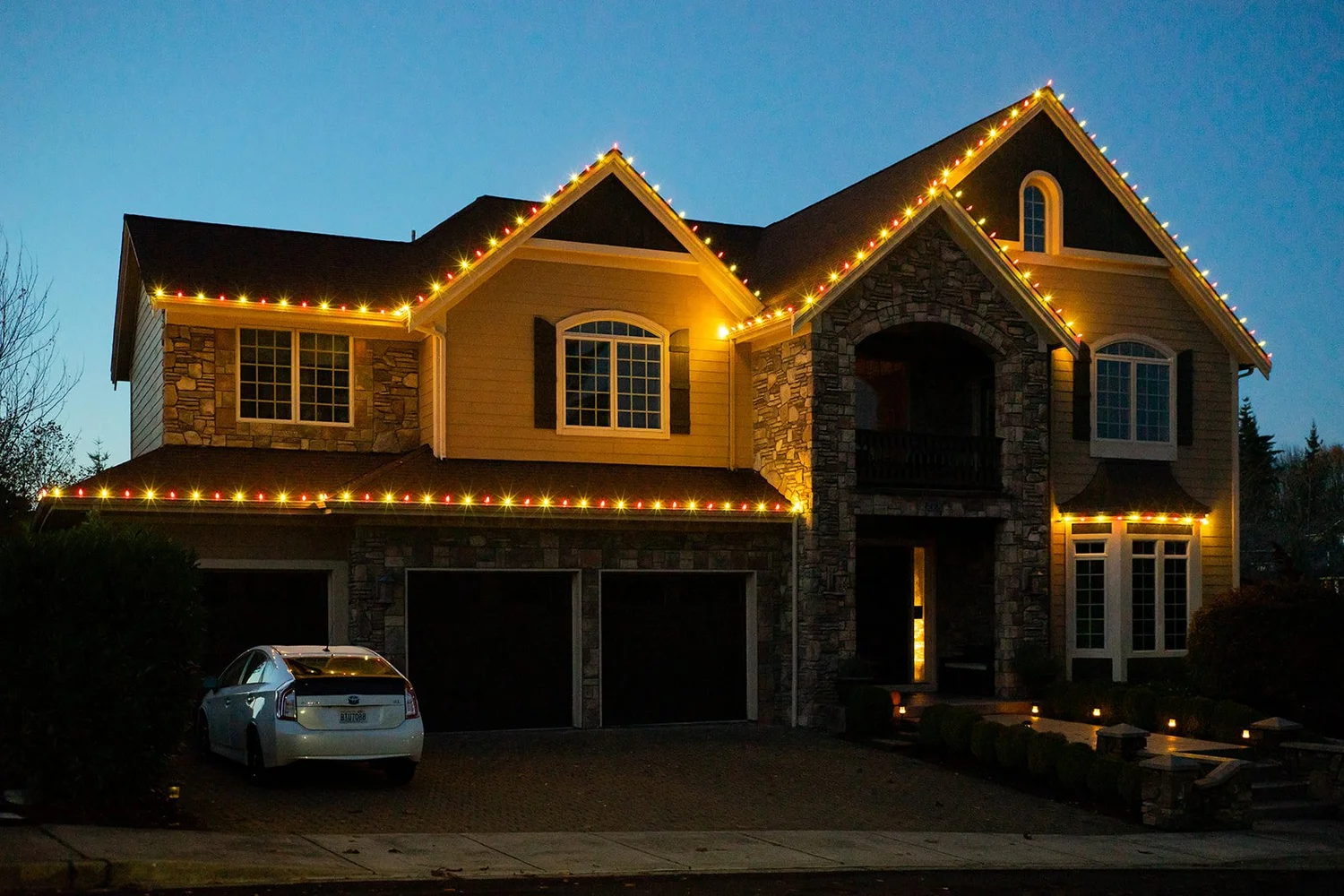 Two-story stone home with warm white C9 Christmas lights along every roofline at dusk