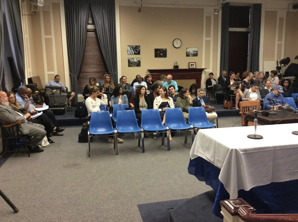 Group of people seated in a room with blue chairs, a table with microphones, and a wall with a clock and framed pictures, ready for a meeting or presentation.