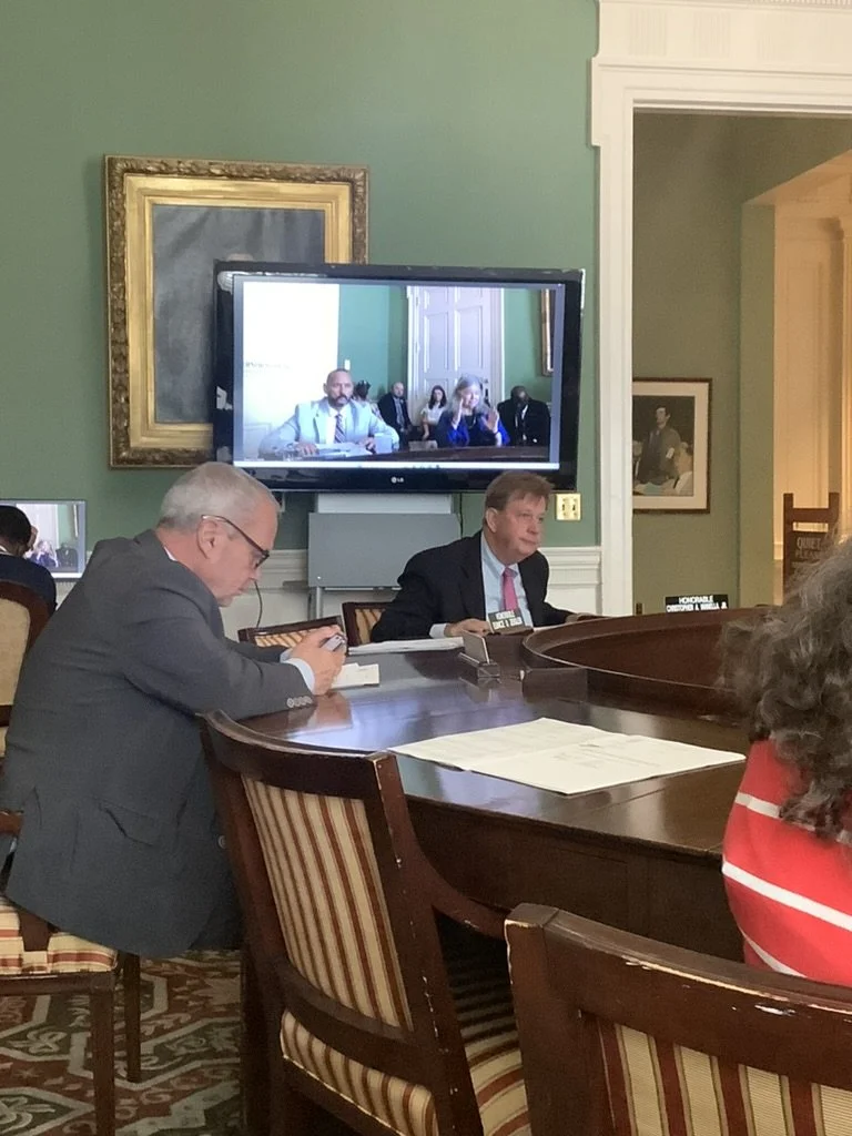 A meeting room with people seated around a large wooden table. One man is using his phone, another man is looking at the camera. A flat-screen TV is mounted on the wall, showing the same room and some attendees.