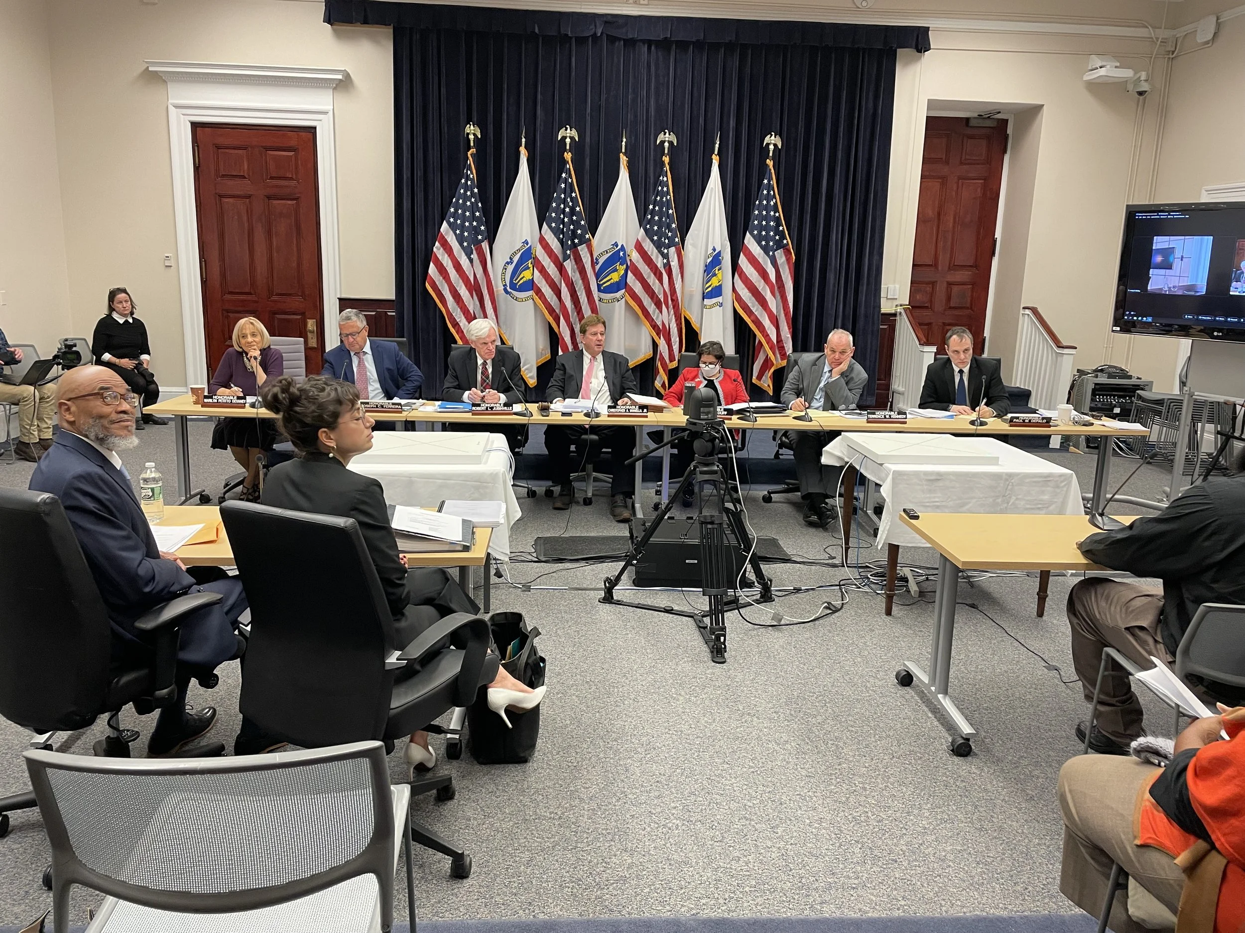 A meeting with several people seated at tables, some taking notes or listening. Eight individuals are seated at a long table in the background with microphones, while others sit around in office chairs. Behind them are seven American flags and govern