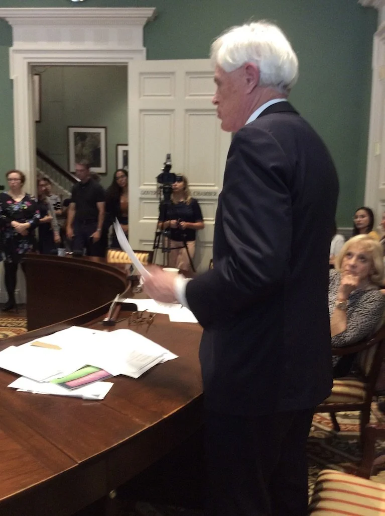 An older man with white hair, dressed in a dark suit, stands at a wooden desk holding papers during a formal event. Several people, including women, are seated and standing around in a room with green walls and white trim. A woman with blonde hair in