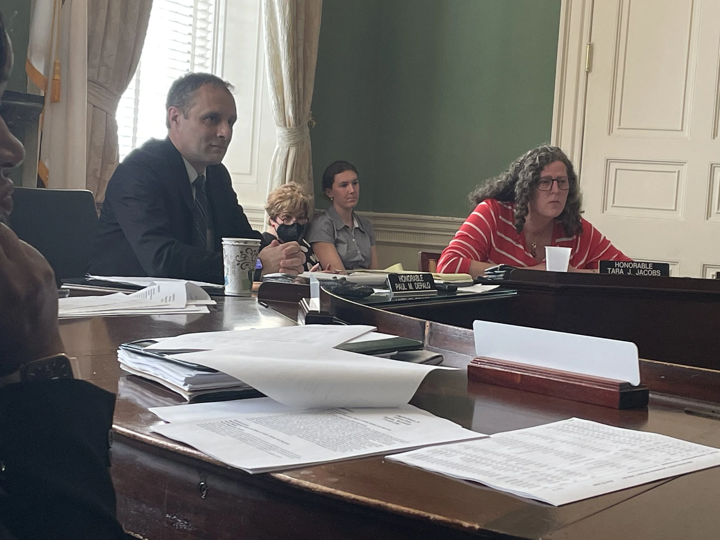 A group of people seated at a conference table in a formal meeting room, with documents, nameplates, and cups in front of them.