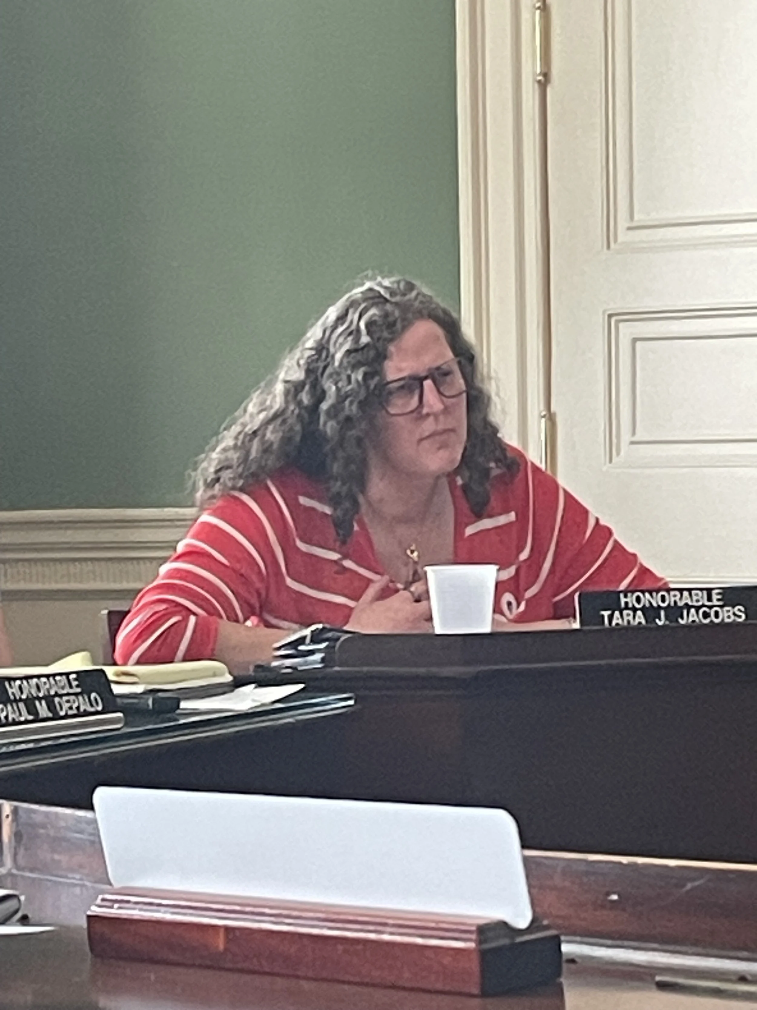 A woman with curly hair, glasses, and a red striped shirt sitting at a table in a formal setting, possibly a government meeting, with a nameplate reading 'Honorable Tara J. Jacobs' and some paperwork and cups on the table.