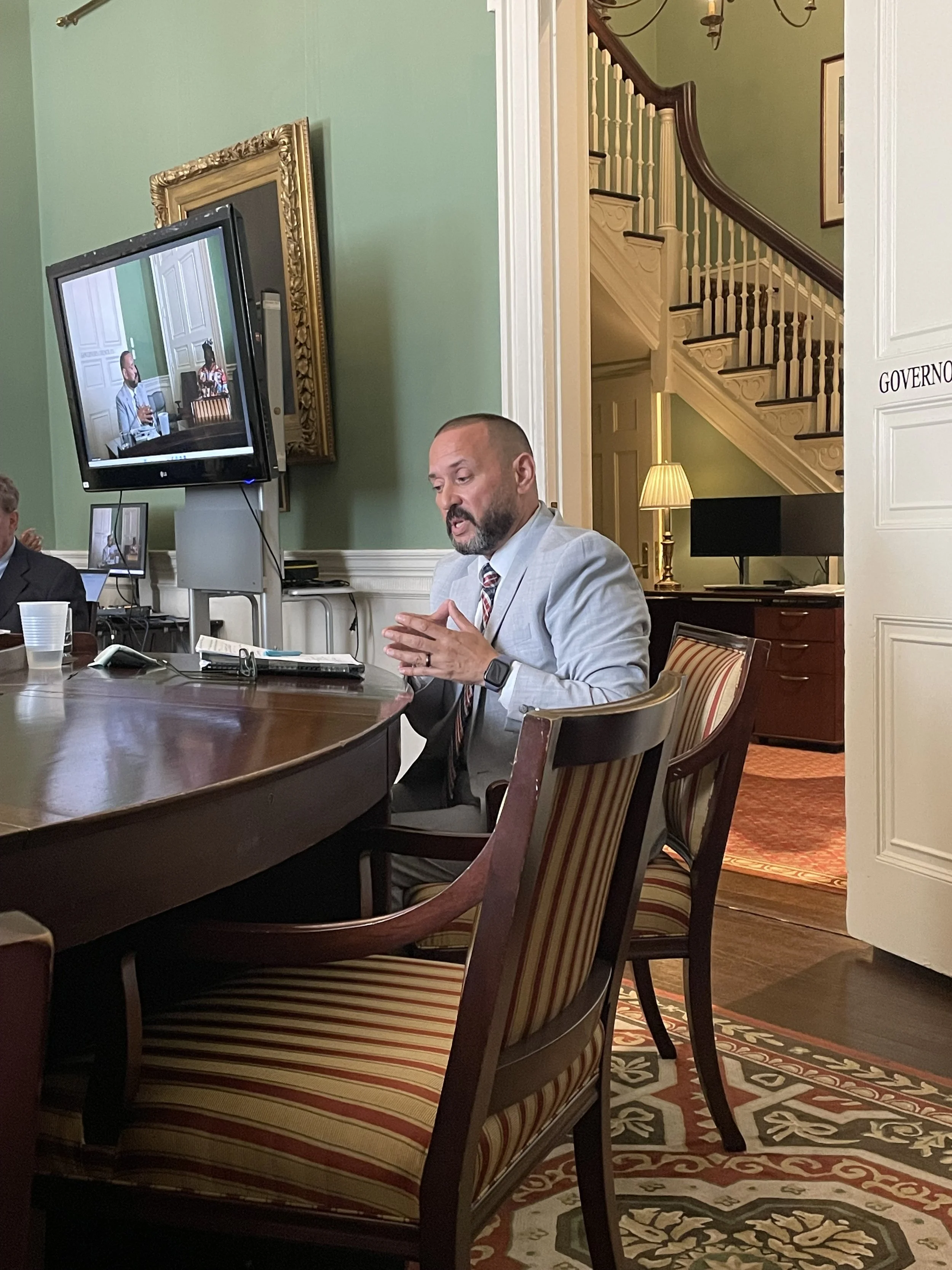 A man in a light gray suit, striped tie, and smartwatch sitting at a conference table, speaking or gesturing, in a formal room with green walls, a staircase, and a television screen behind him showing a similar scene.