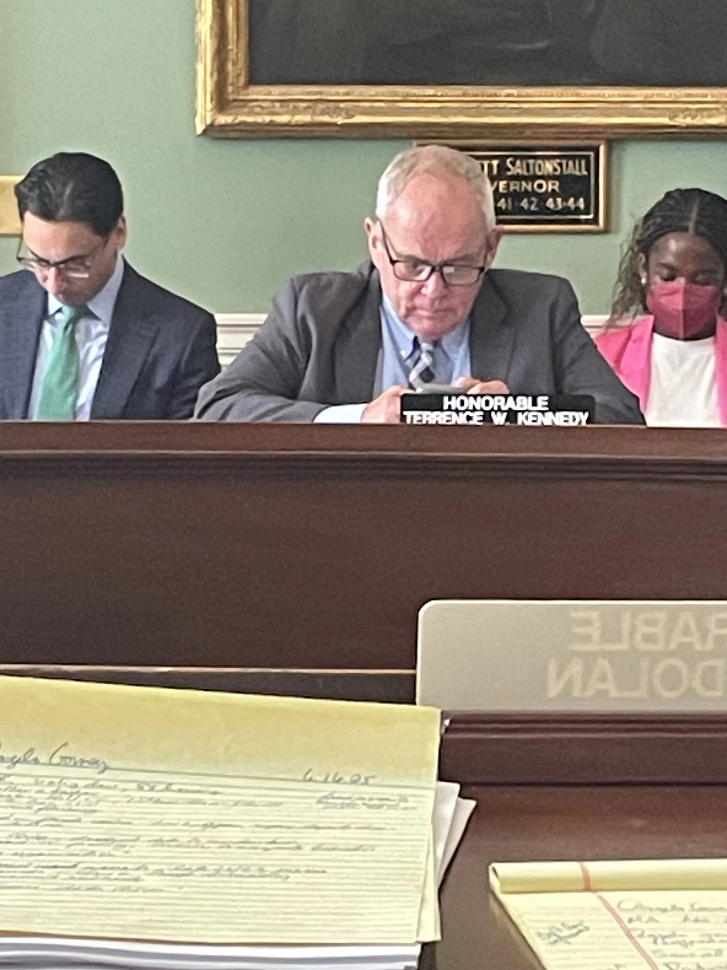 A formal meeting room with three people sitting at a desk with a sign reading 'Honorable Terence W. Kennedy' in front of the man in the middle. The man on the left is looking down, the man in the middle is looking at his phone, and the woman on the r