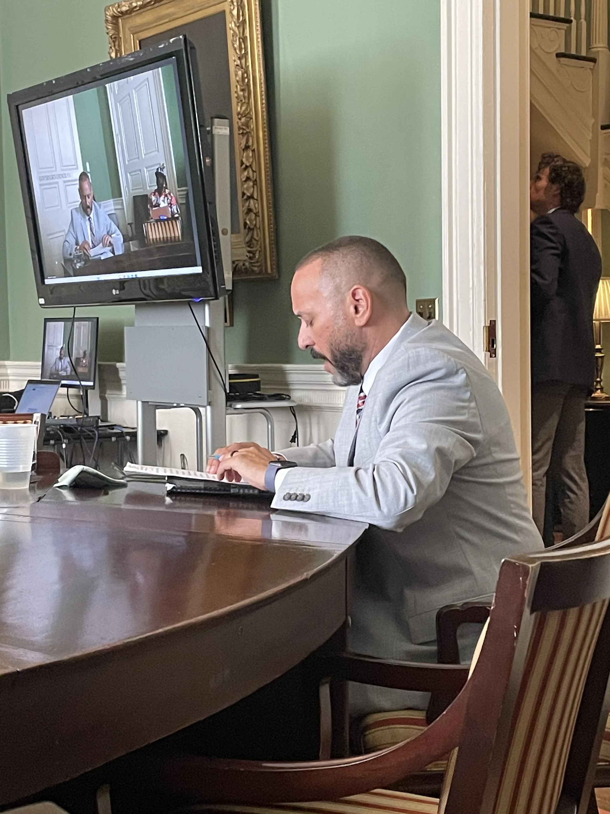 A man in a light gray suit sits at a polished wooden conference table, working on a laptop. He is in a room with green walls, decorated with a gold-framed mirror. A large monitor on a stand displays a video conference with two other people, one man a