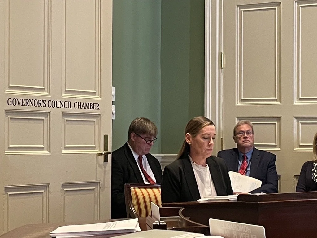 Three individuals in business attire sitting at a table in the Governor's Council Chamber, with a sign on the door reading 'Governor's Council Chamber'.