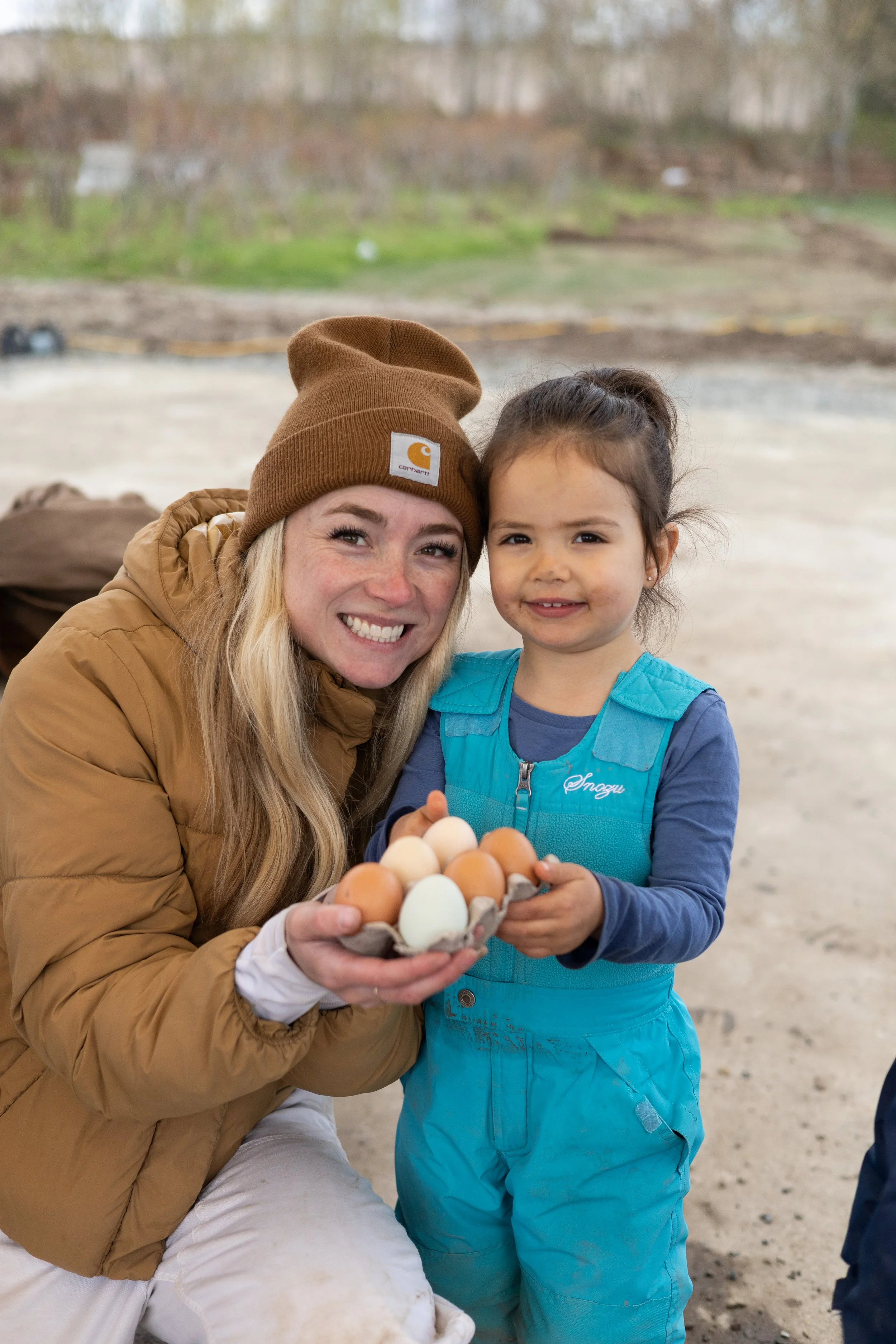 A smiling woman holding a tray of assorted eggs, standing next to a young girl holding the tray, outdoors with a blurred background of trees and ground.