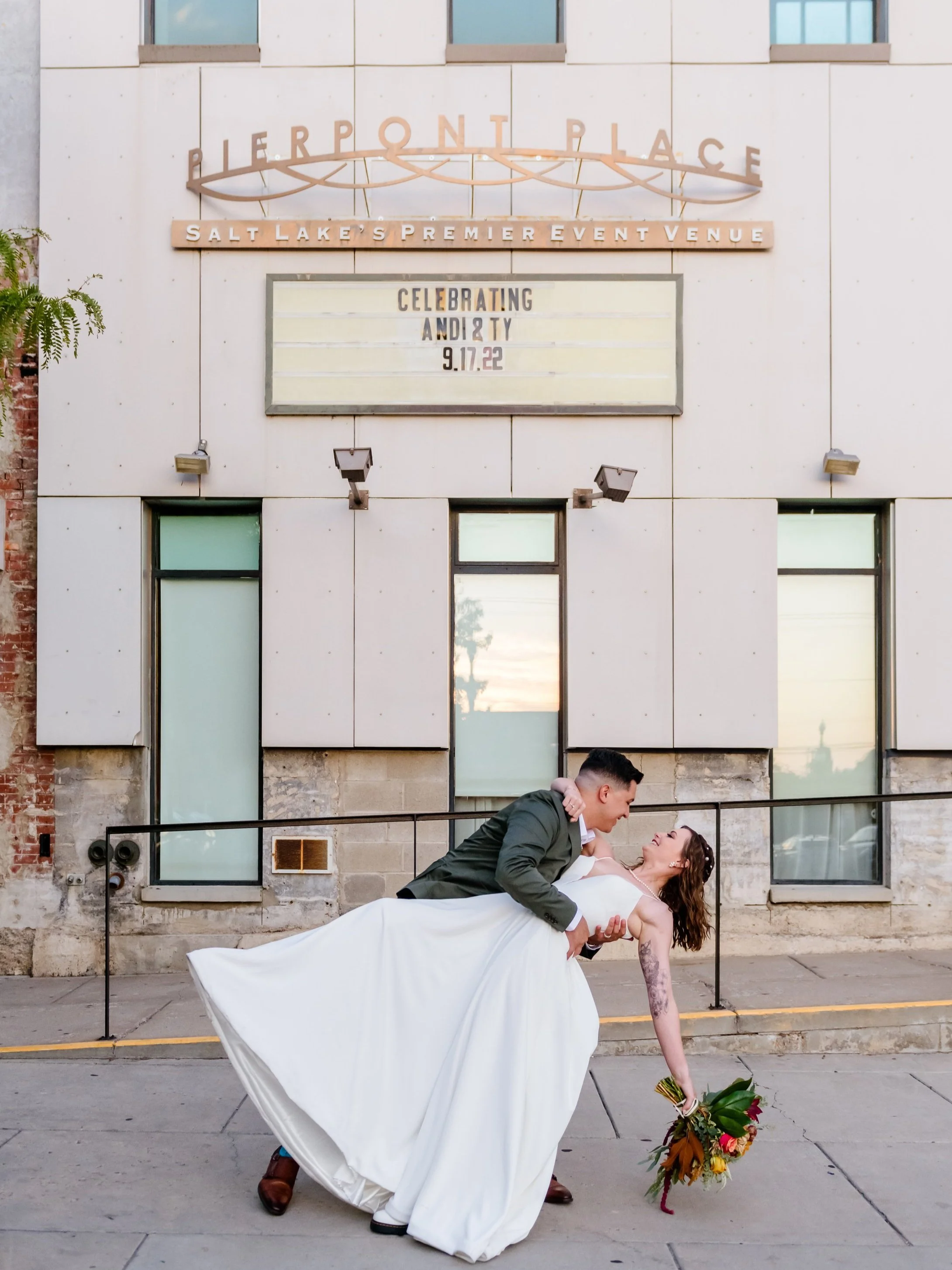 A newlywed couple dancing outside Pierpont Place, a Salt Lake City event venue, with the groom in a dark green jacket and the bride in a white wedding dress, holding a bouquet of flowers.