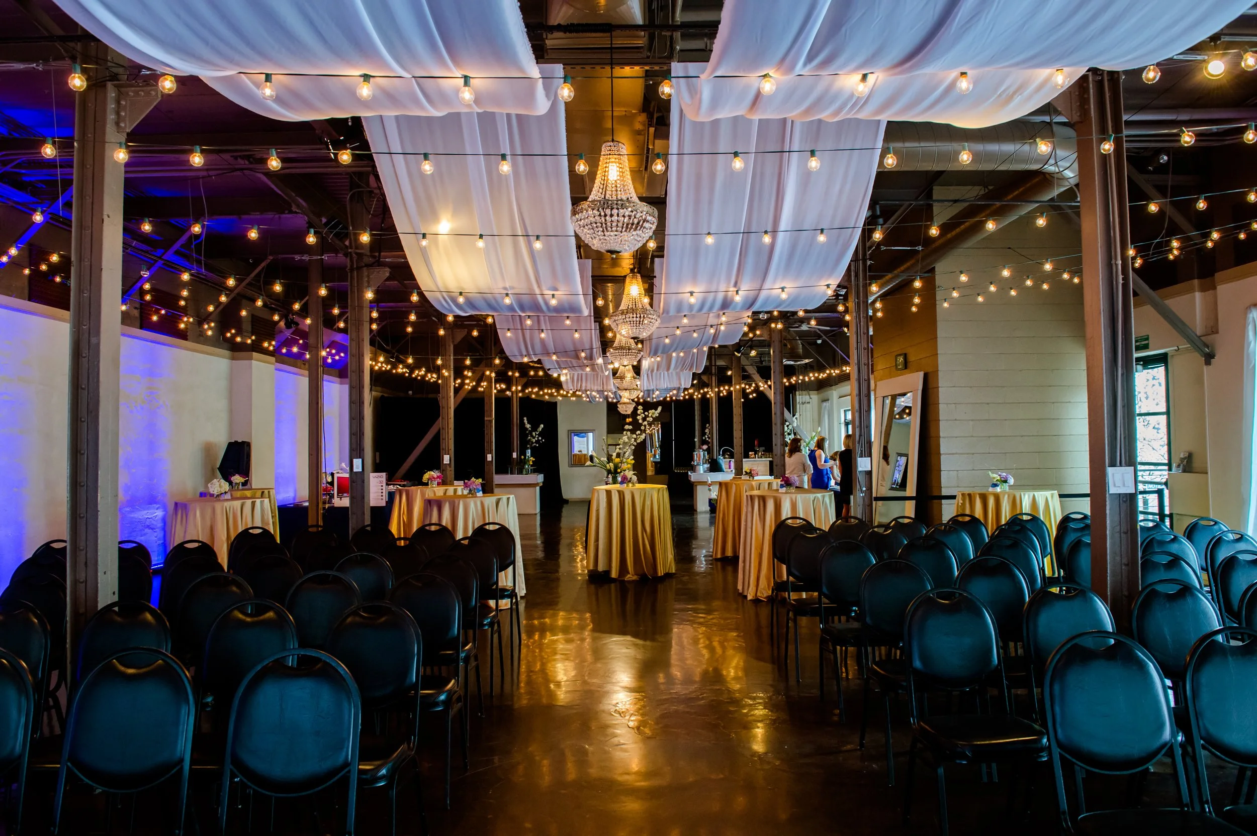Wedding reception area with rows of black chairs, round tables with yellow tablecloths, hanging string lights, and chandeliers, in a decorated indoor venue.