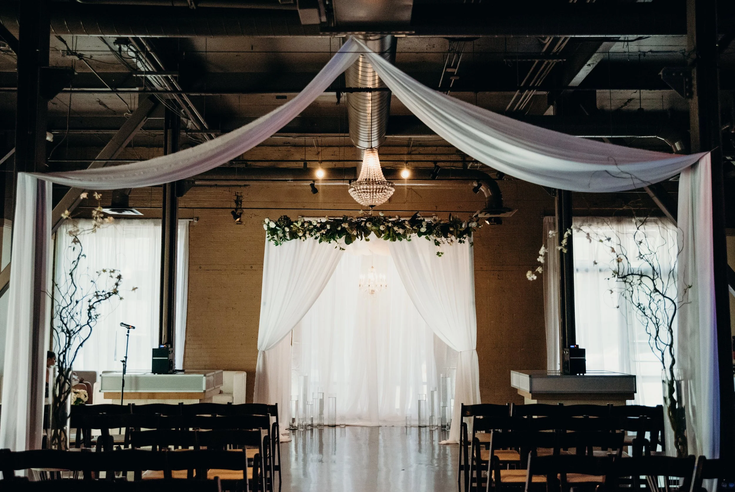 An indoor wedding ceremony setup with white drapes, floral garland, chandelier, and chairs facing the altar.
