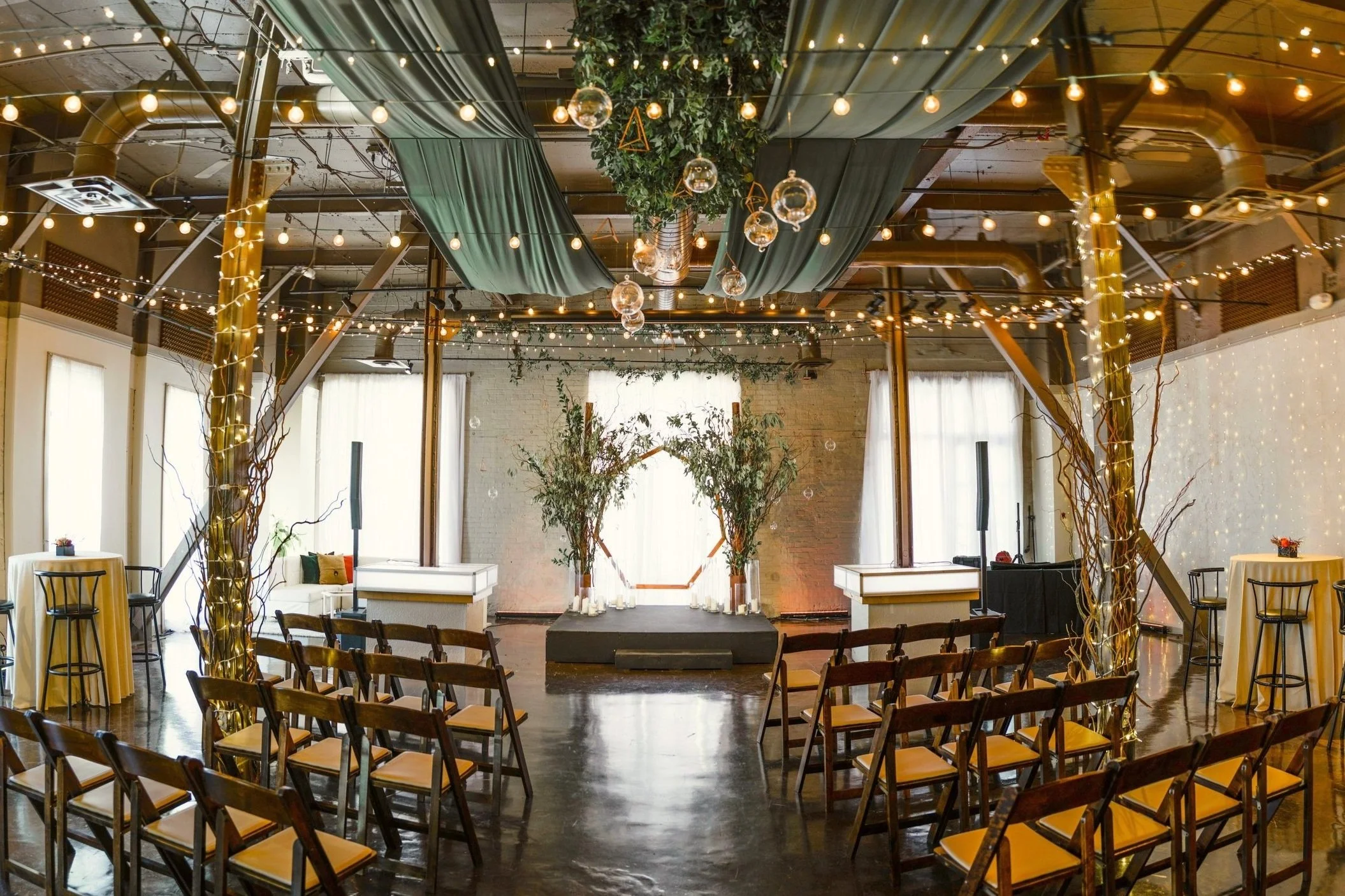Indoor wedding or event venue decorated with fairy lights, hanging glass orbs, lush greenery, and rustic chairs arranged facing a small stage with tall floral arrangements.