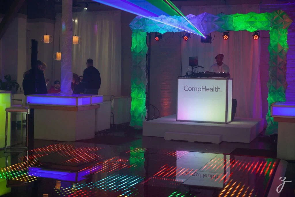 Indoor event space with colorful lighting, a DJ on stage, glowing dance floor, and a few people socializing.