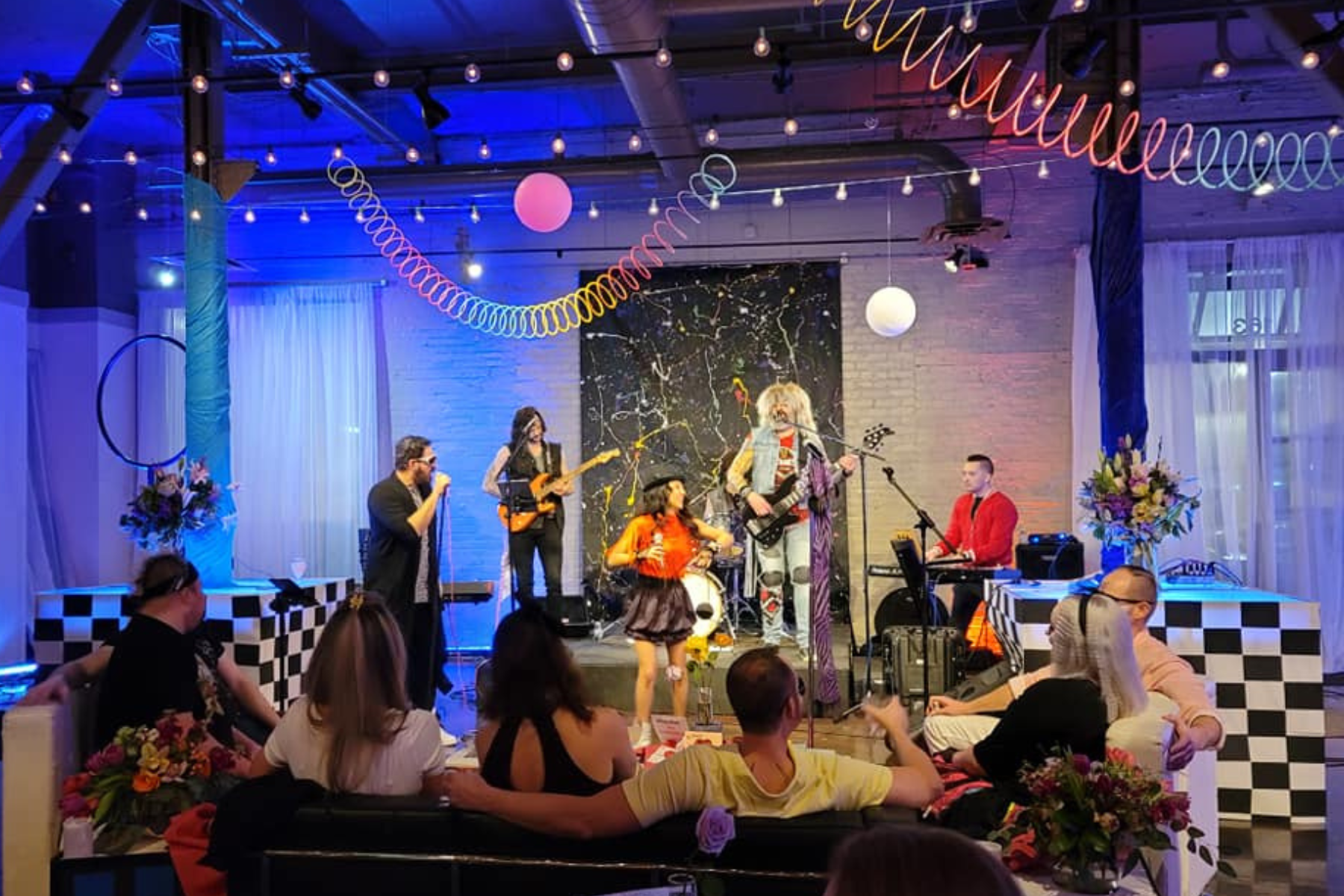 Live band performing on stage at a colorful party or event, with decorations including string lights, paper lanterns, and floral arrangements, and guests seated watching the performance.
