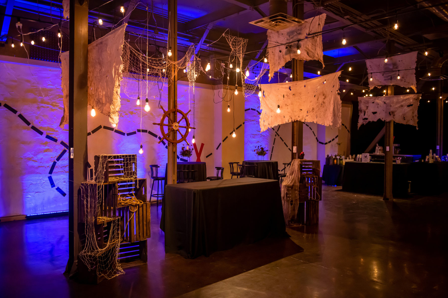 A dimly lit indoor event space decorated with pirate-themed decorations, including tattered cloths hanging from the ceiling, a ship's wheel on the wall, and string lights creating a moody ambiance.