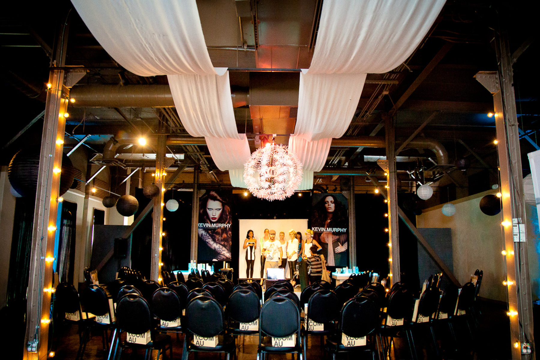 Indoor event space with a stage, black chairs arranged in rows, and large posters of models. There are hanging white and black paper lanterns, and a white chandelier. The ceiling has white fabric drapes and industrial piping.