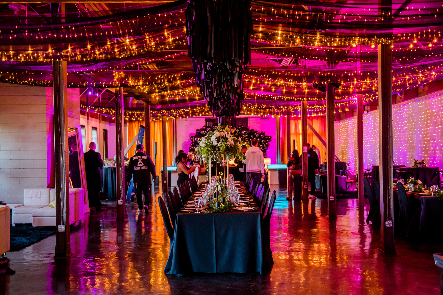 Decorated event venue with string lights on the ceiling, arranged dining tables with floral centerpieces, and people preparing for the event.