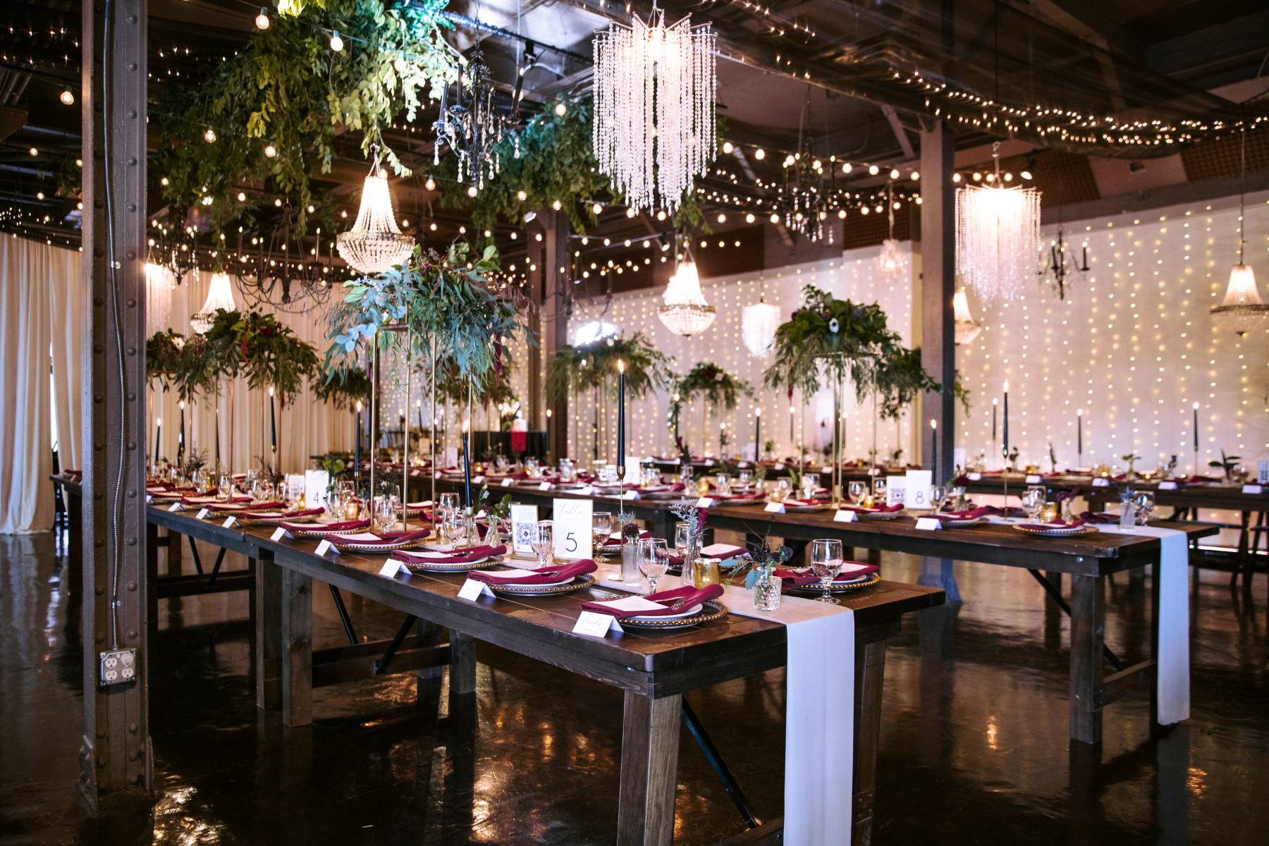 Long wedding reception tables decorated with floral centerpieces, candles, and table settings, illuminated by chandeliers and string lights in a rustic indoor venue.
