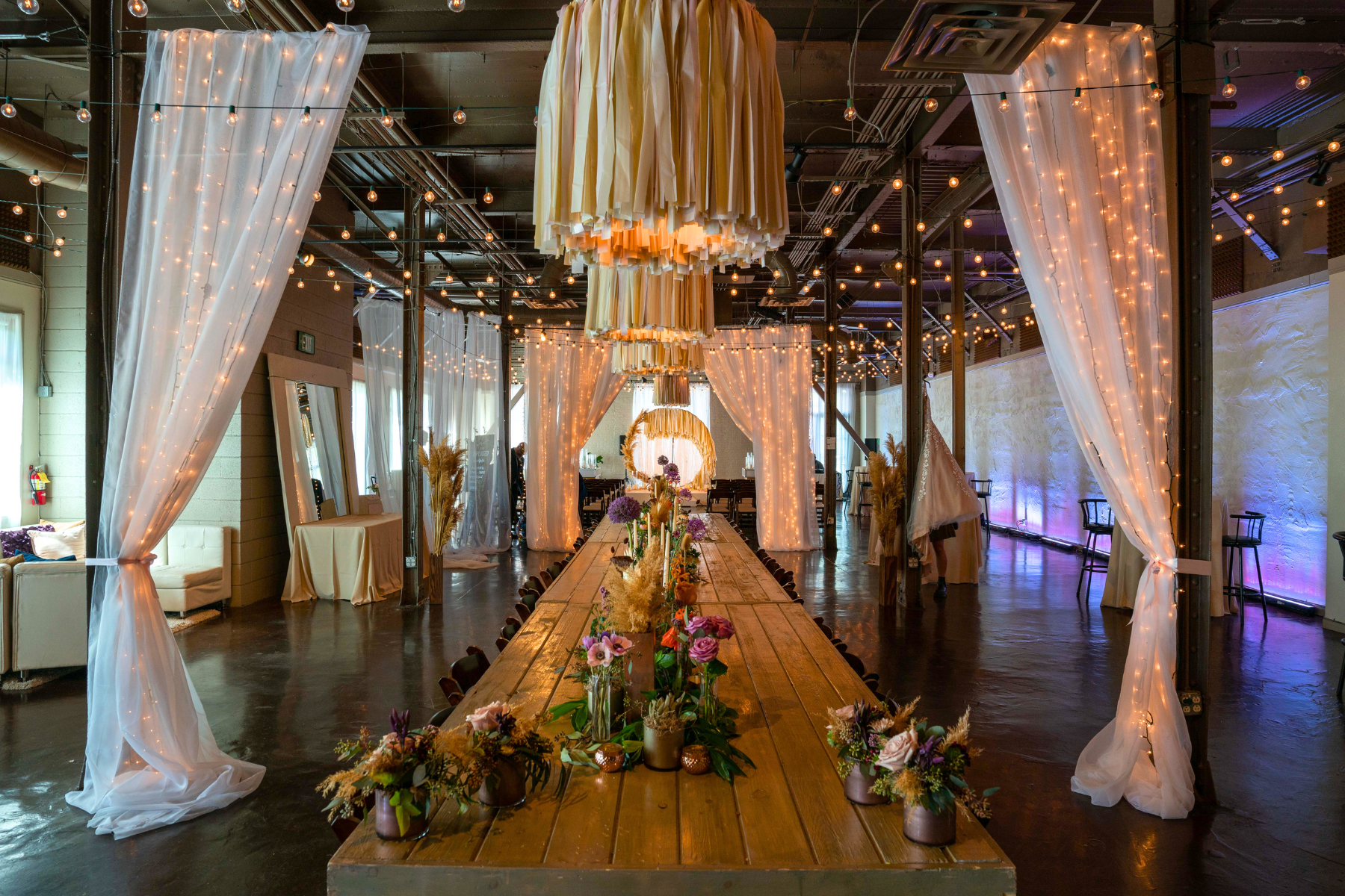Decorated event space with long wooden table, flowers, string lights, curtains, and hanging fabric chandelier