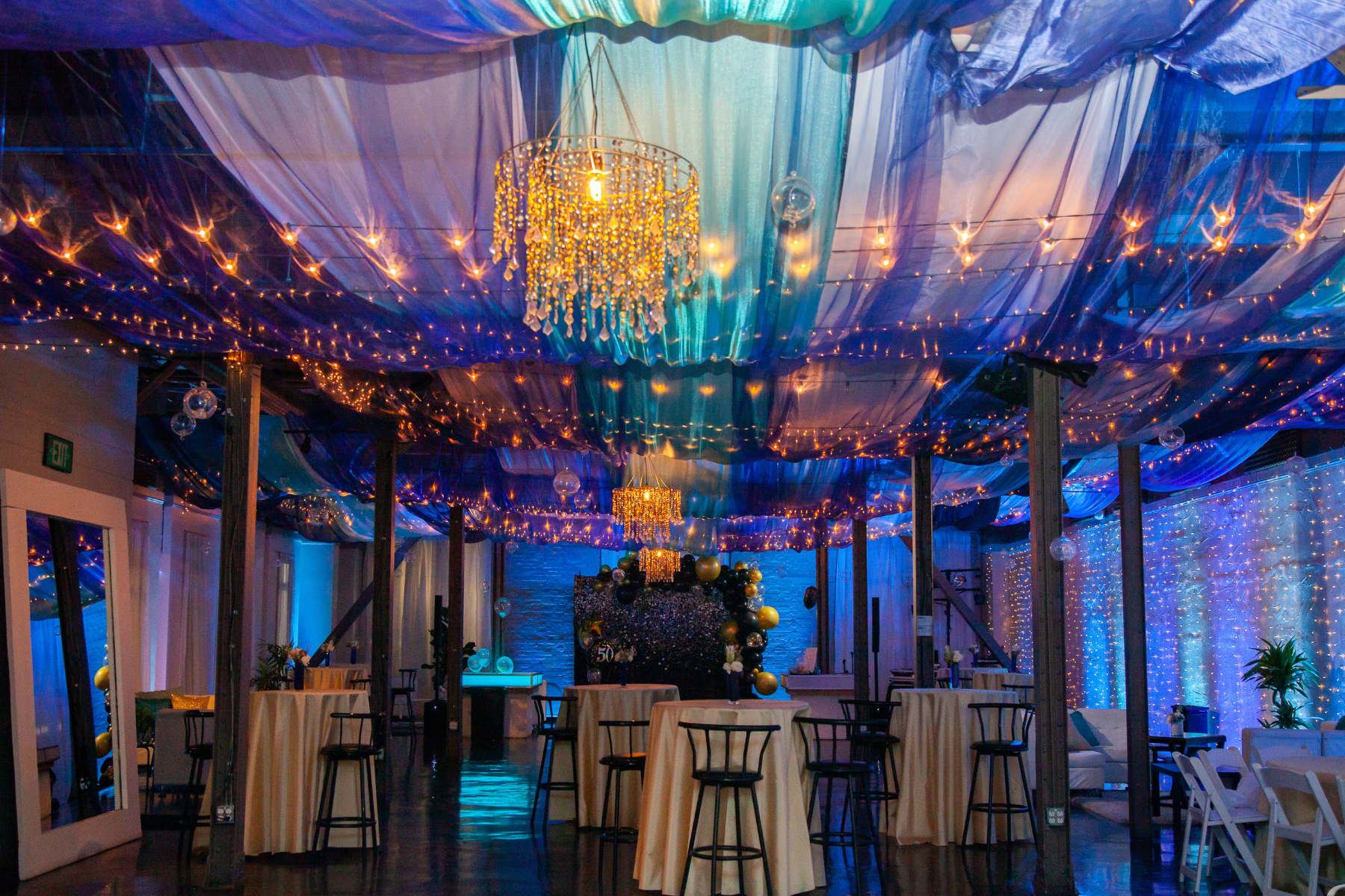 Decorated event space with blue and purple draped fabric on the ceiling, string lights, hanging glass globes, and a central chandelier with beads. There are high tables with yellow tablecloths and black chairs, and a backdrop with balloons and floral