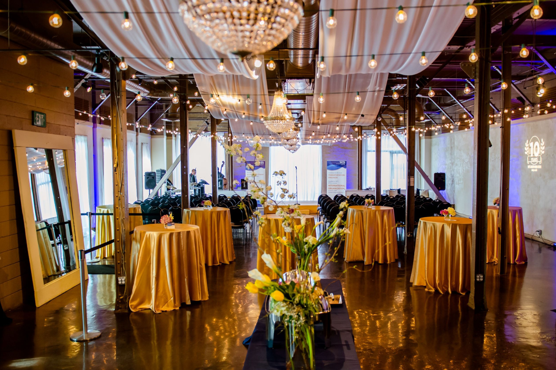 Elegant event space with round high-top tables draped in gold tablecloths, floral centerpieces, string lights and chandeliers hanging from the ceiling, and a stage with two people speaking in the background.