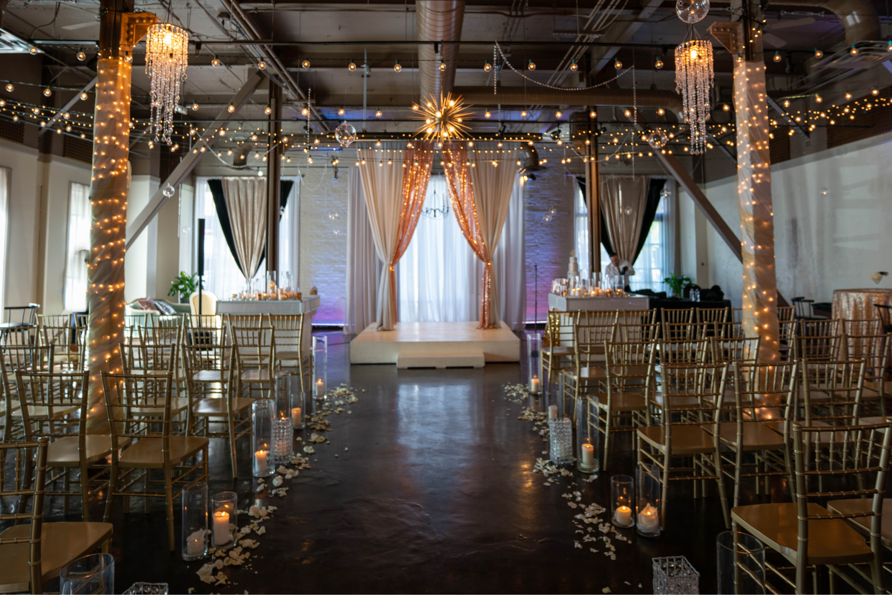 Elegant wedding ceremony setup with a small stage, draped curtains, decorative lighting, candle centerpieces along the aisle, and rows of gold chairs.