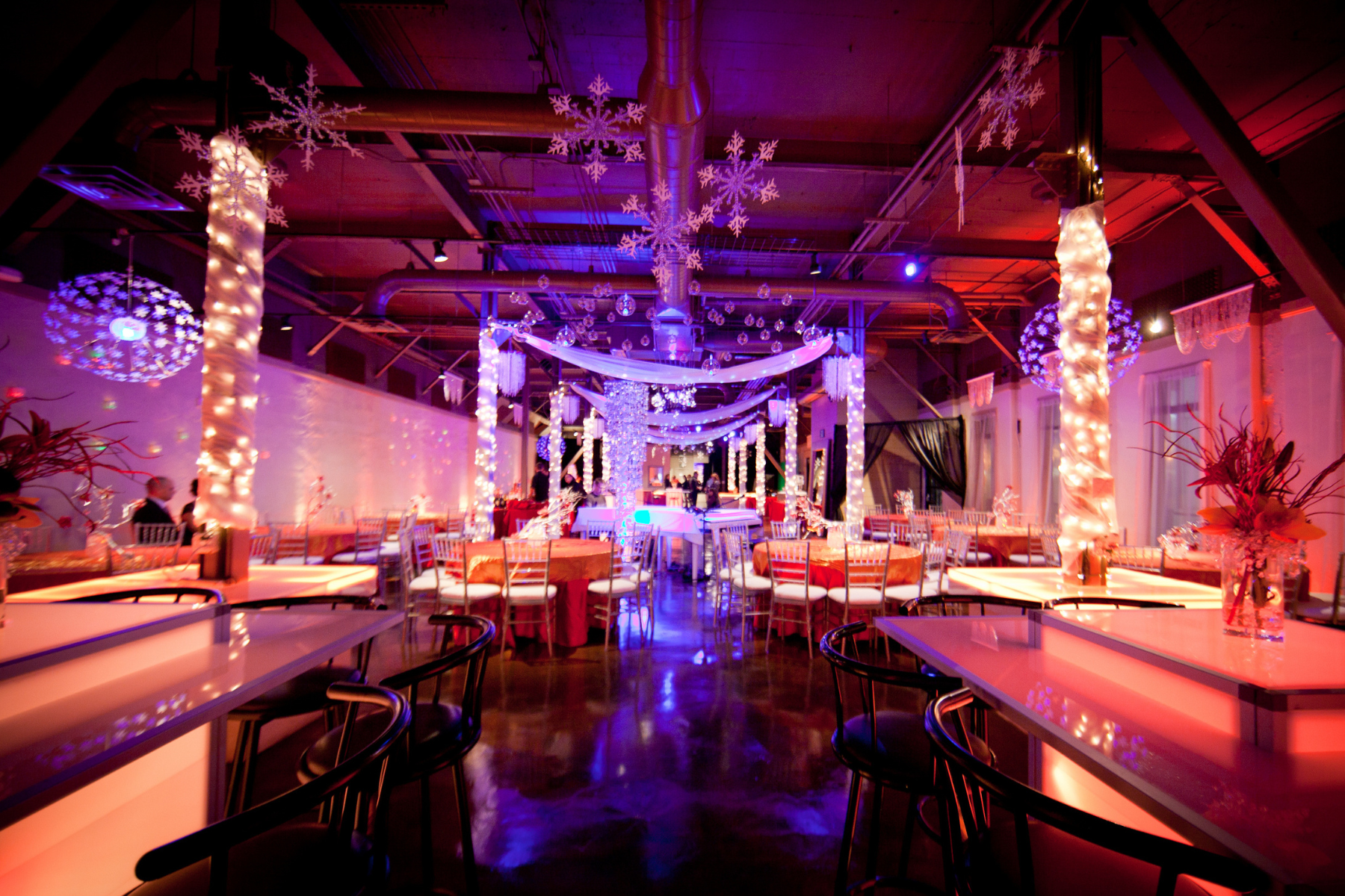 A decorated event space with tables, hung drapes, and snowflake decorations, illuminated by purple and pink lights.