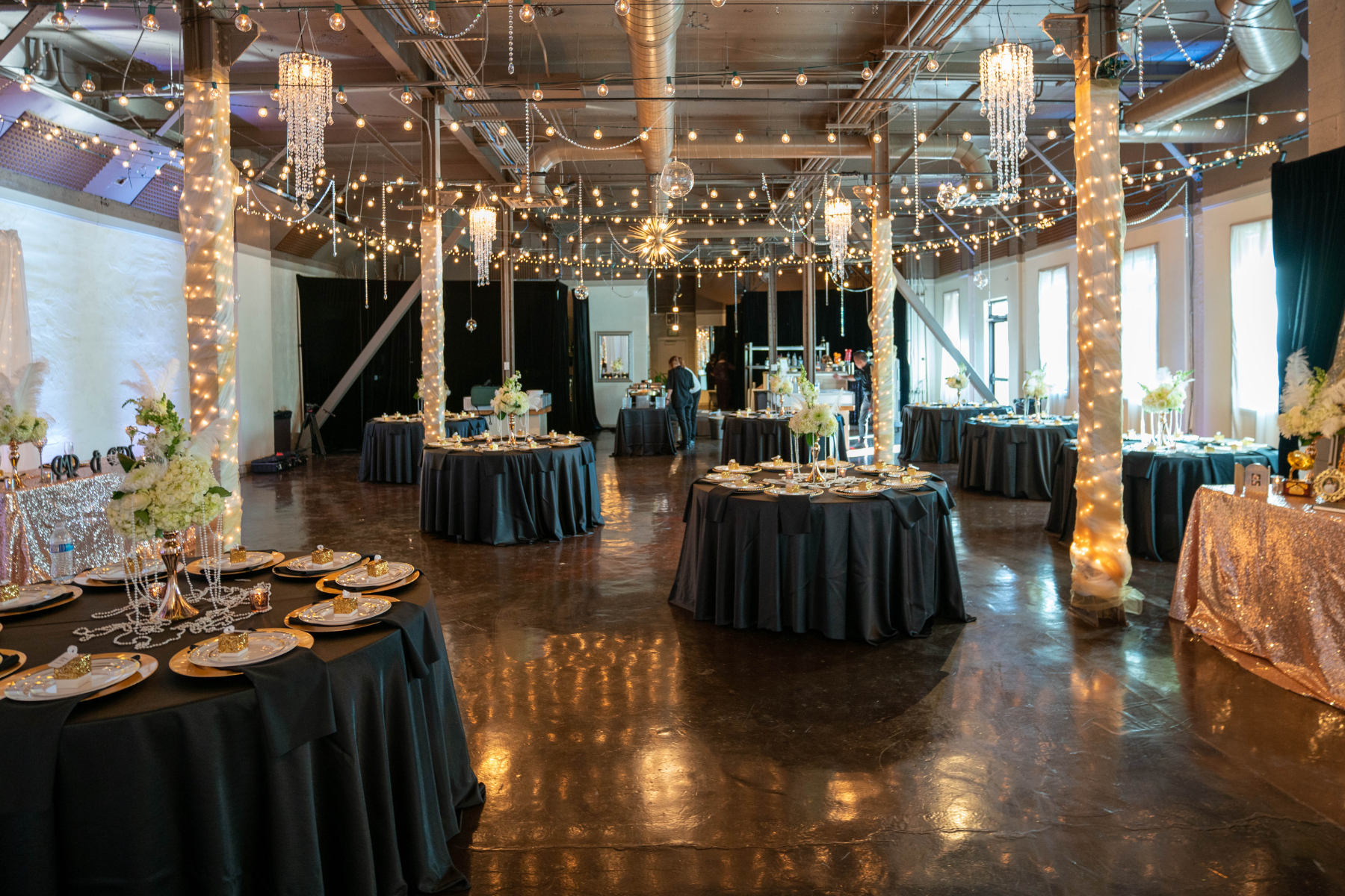 Elegant event space decorated with fairy lights, chandeliers, and floral centerpieces on round tables with black tablecloths, set for a celebration such as a wedding or party.