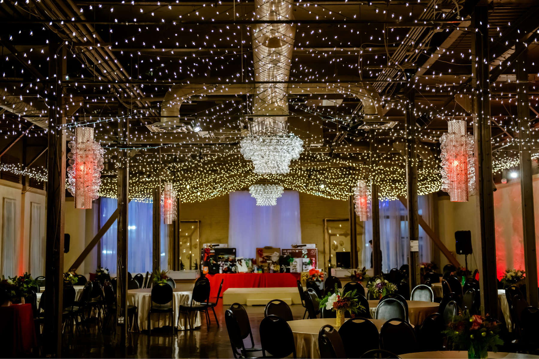 A decorated event space with hanging fairy lights and chandeliers, round tables covered with beige and red tablecloths, chairs, and floral centerpieces, set up for a celebration or banquet.