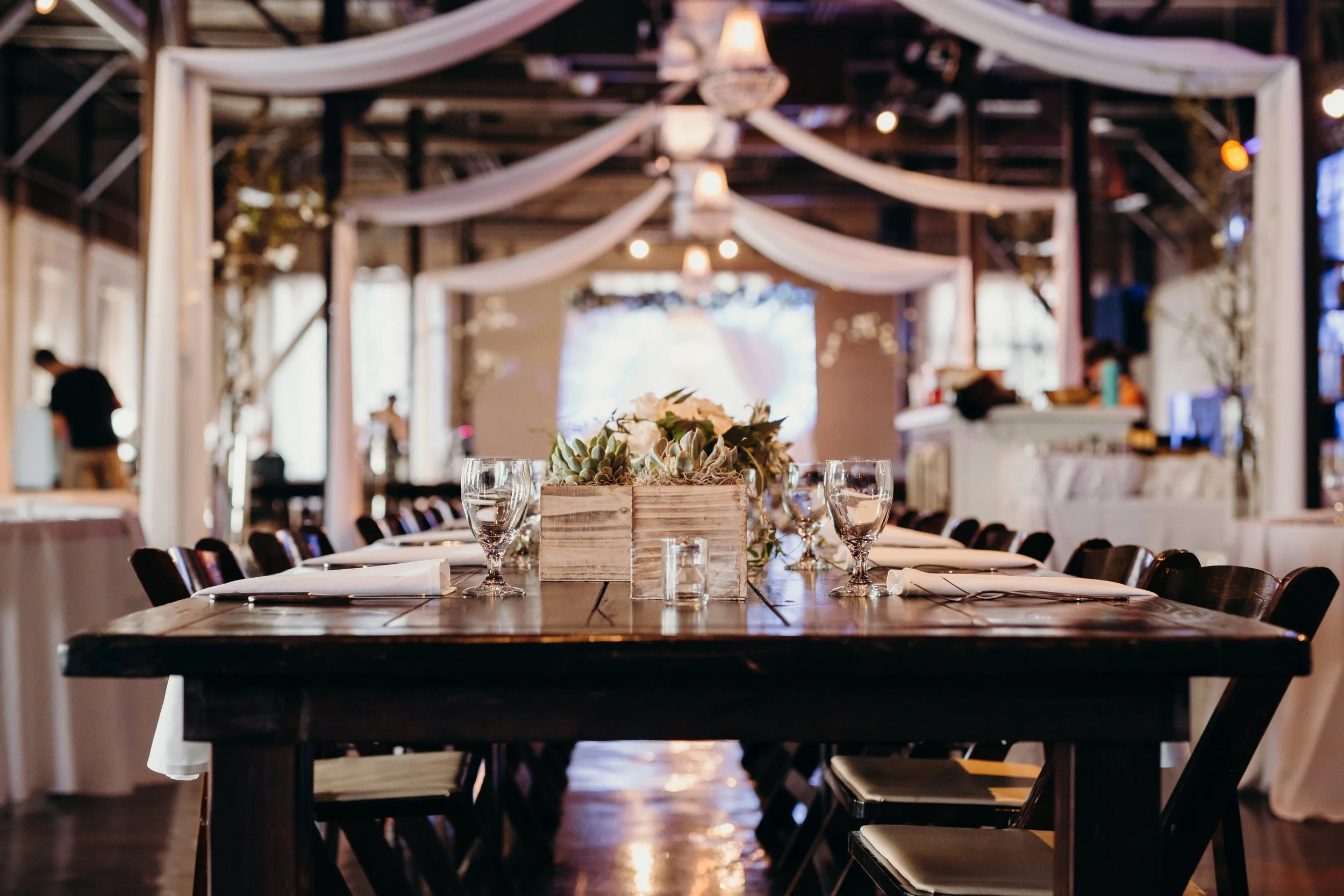 A decorated wedding reception table with glasses and napkins, a floral centerpiece, and a rustic wood table in a barn-style venue with draped white fabric and chandeliers.