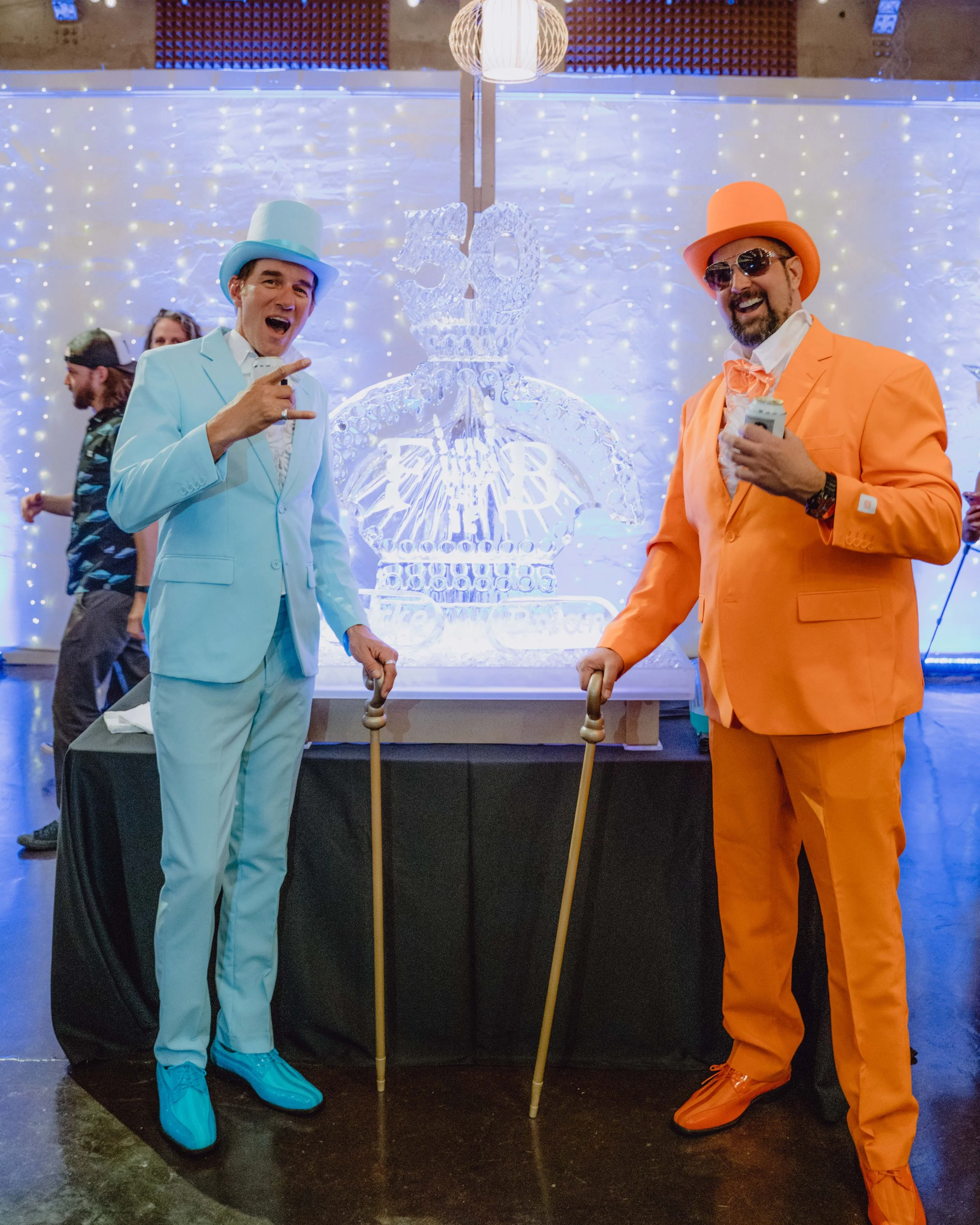 Two men dressed in colorful suits and hats, one in a light blue suit with a matching top hat and the other in an orange suit with an orange top hat, standing in front of an ice sculpture of a bottle with a crown and the number '50' on it. They are holding canes and smiling, at a celebration event with a decorated background and other guests in the background.