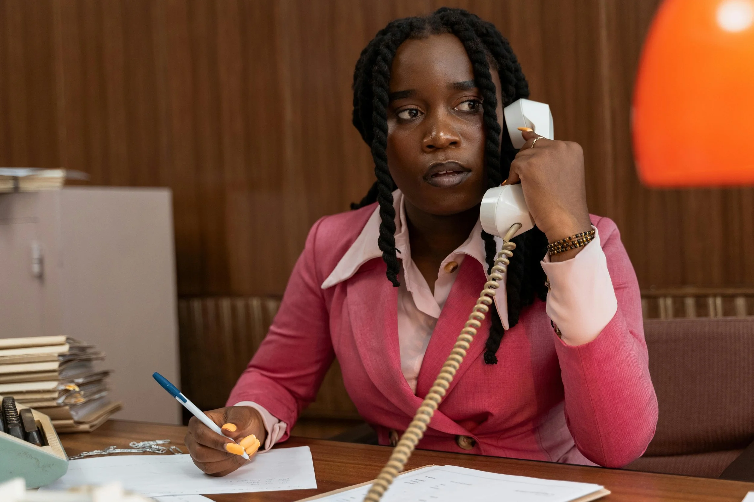 A woman with braided hair talking on a landline phone at her desk in an office, holding a pen over papers.