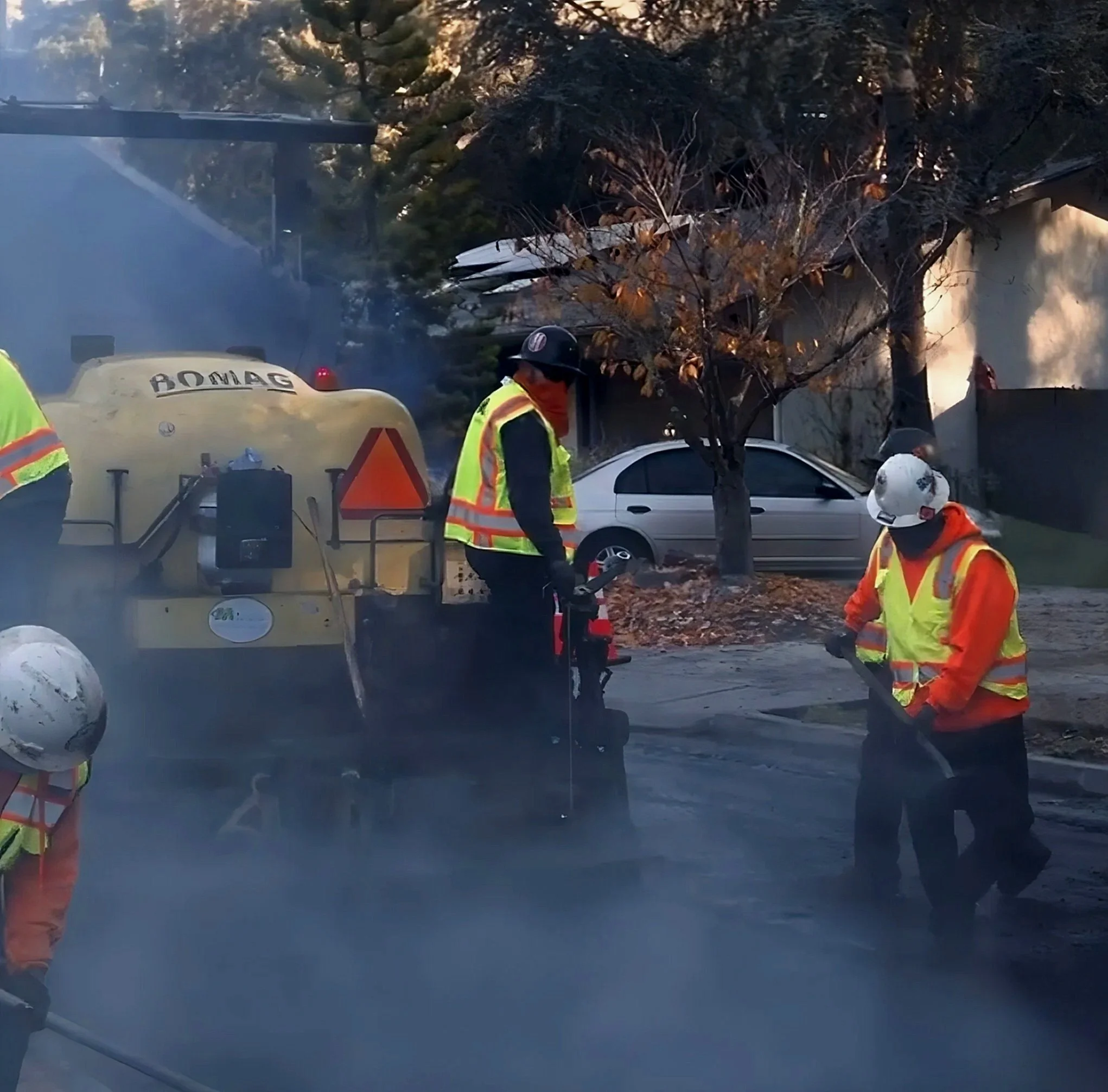 Firefighters work on a street with smoke in the air, near a house with a burning tree, using tools and equipment.