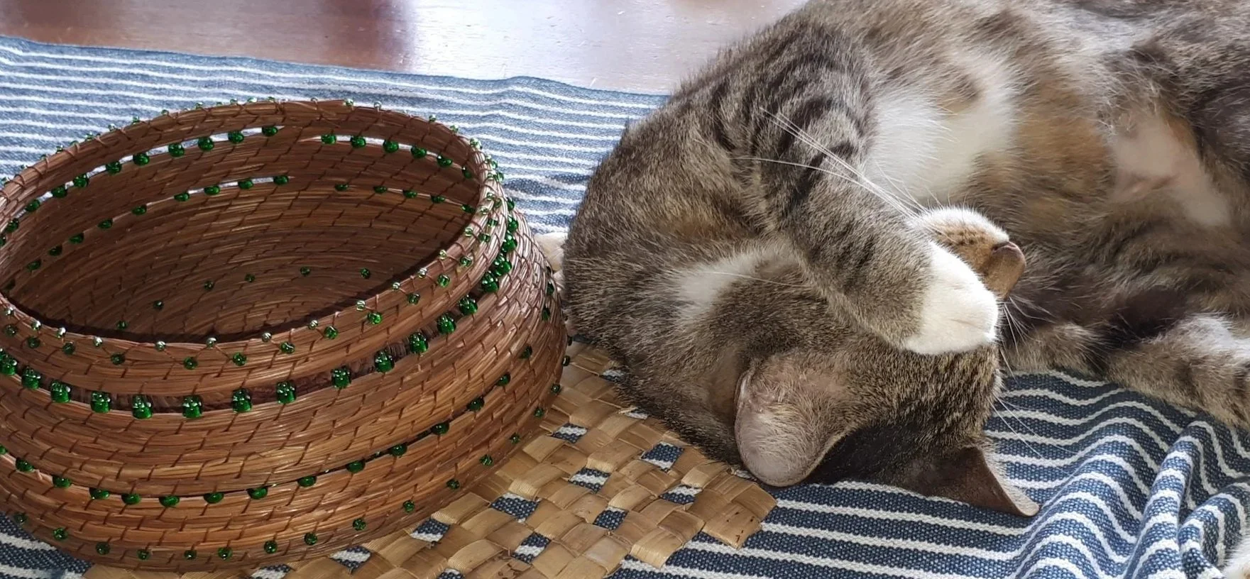 A tabby cat with white paws lying on a blue striped cloth, next to a woven basket decorated with green beads.