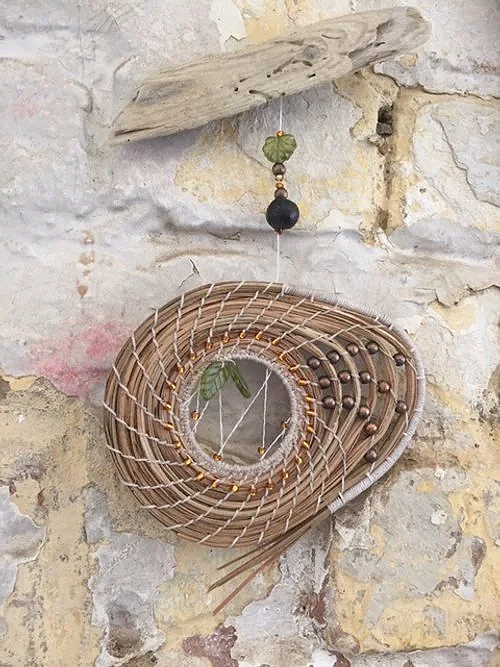 A decorative wall hanging with a coiled pine needle spiral and a hanging beaded ornament against a textured stone wall.