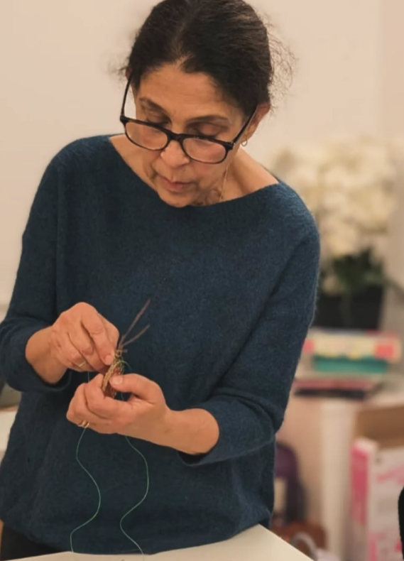 A woman with dark hair, glasses, and a blue sweater, focused on knitting with green yarn.