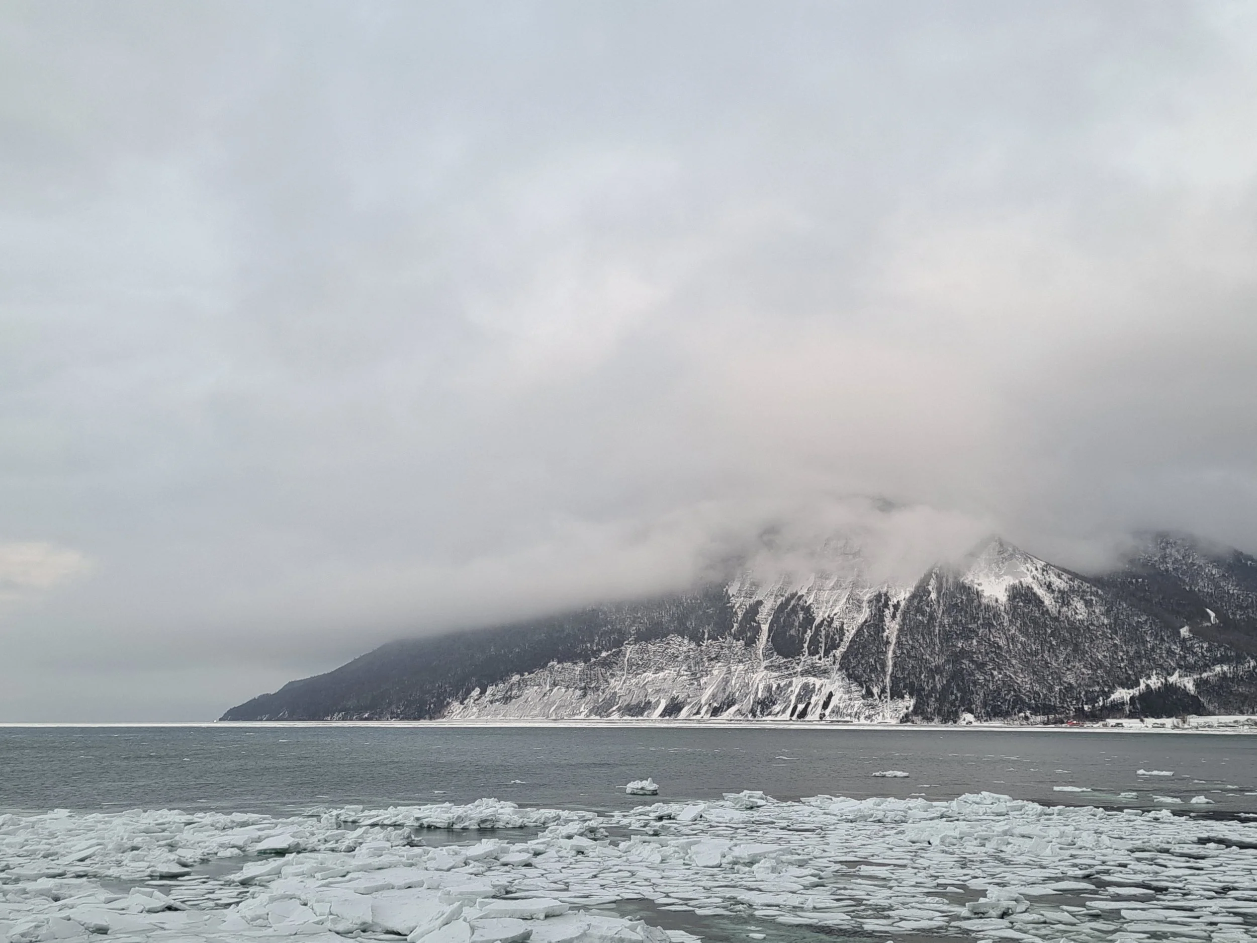 Snow-covered mountains partially obscured by clouds and fog, with ice floating on the water in a cold, winter landscape.