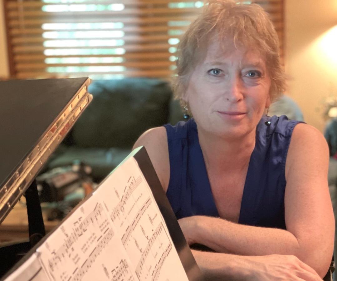 A woman with short blonde hair and blue eyes, wearing a sleeveless blue top, sitting at a piano with sheet music, in a cozy room with wooden blinds and warm lighting.