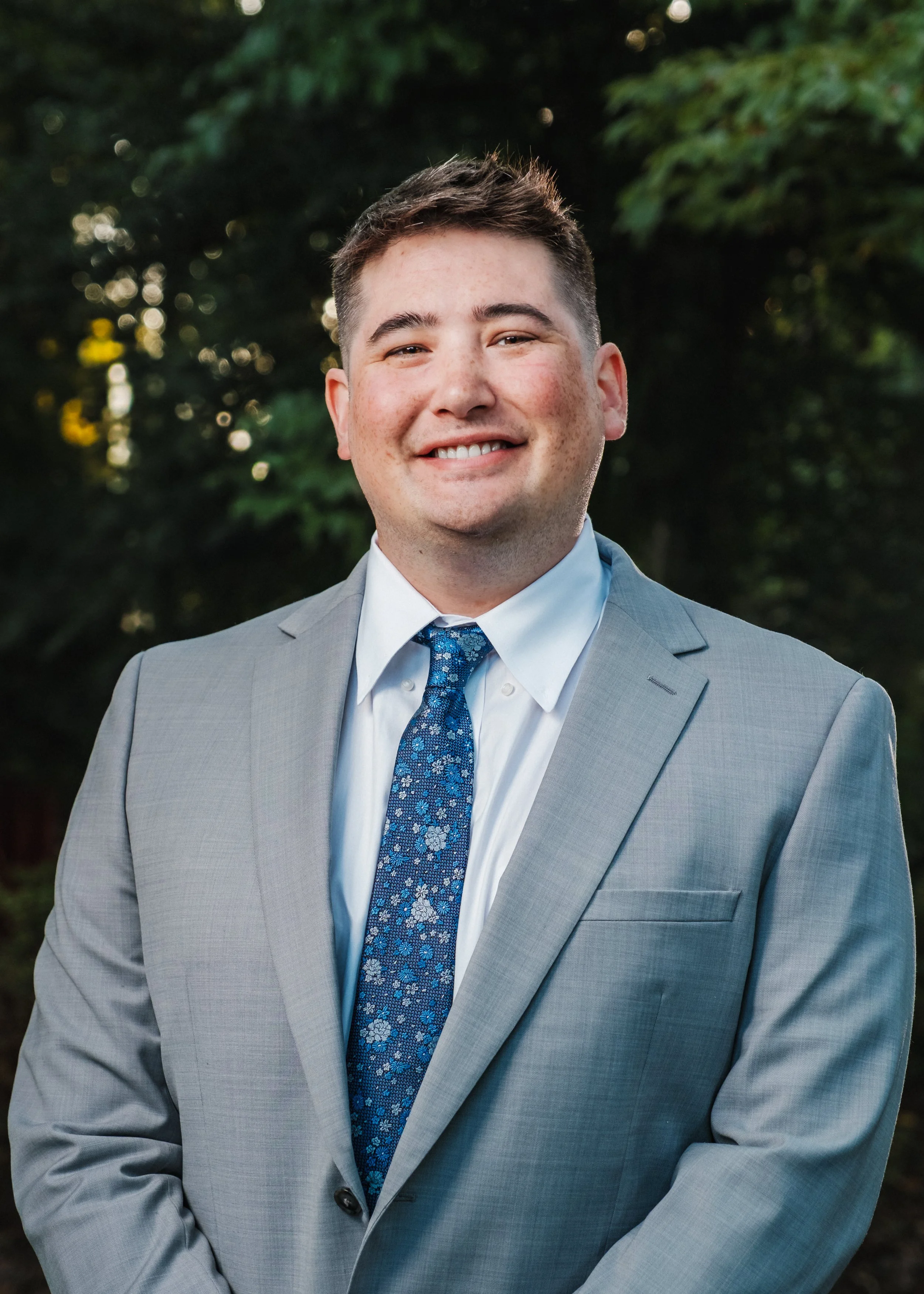 A young man in a light gray suit, white shirt, and blue floral tie, smiling outdoors with trees in the background.