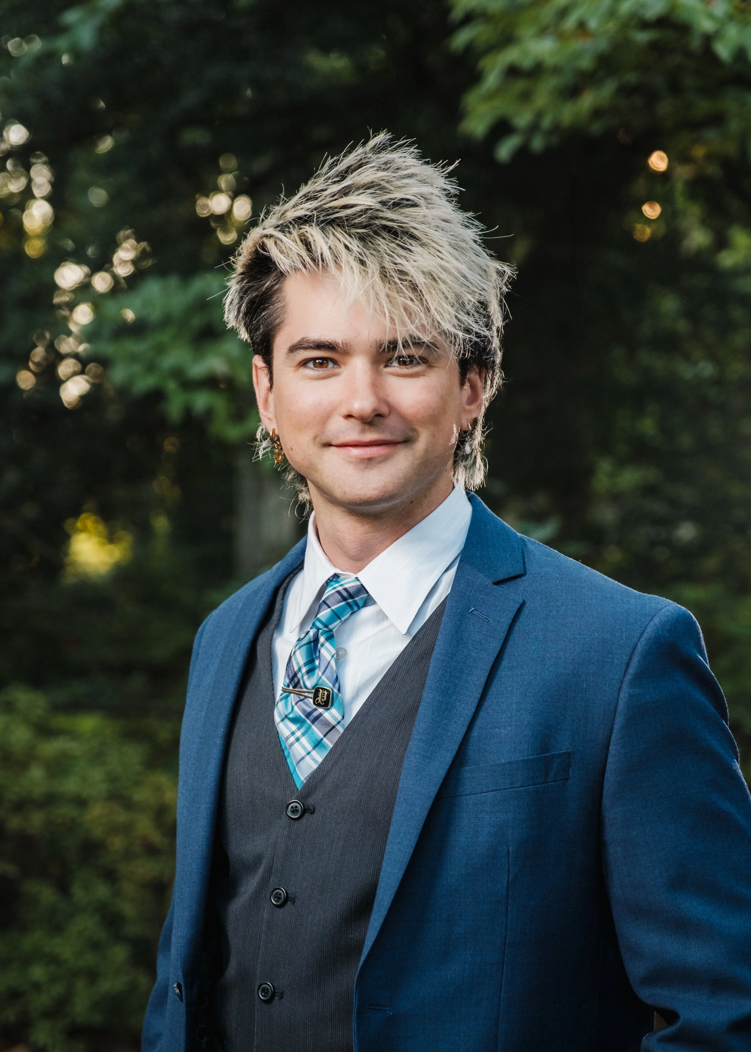 A young man in a dark blue suit with a gray vest, white shirt, and blue plaid tie, standing outdoors in front of greenery and smiling at the camera.